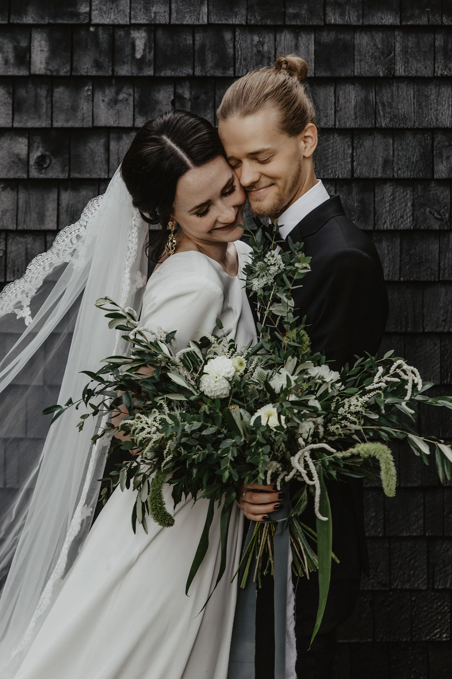 Ett brudpar omfamnar varandra och ler med stängda ögon framför en mörk trävägg i Sävsjö under en bröllopsfotografering. 