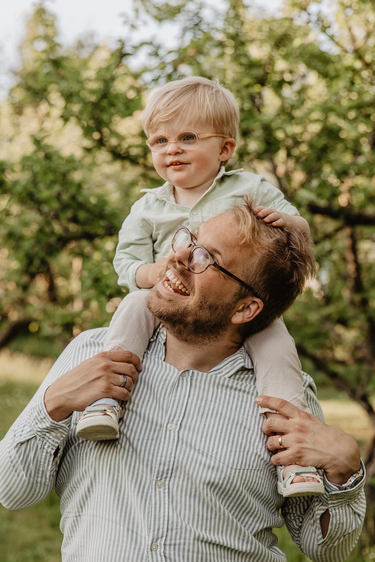 En vuxen man bär en liten barn på sina axlar utomhus, båda är glada och skrattar, med gröna träd i bakgrunden.