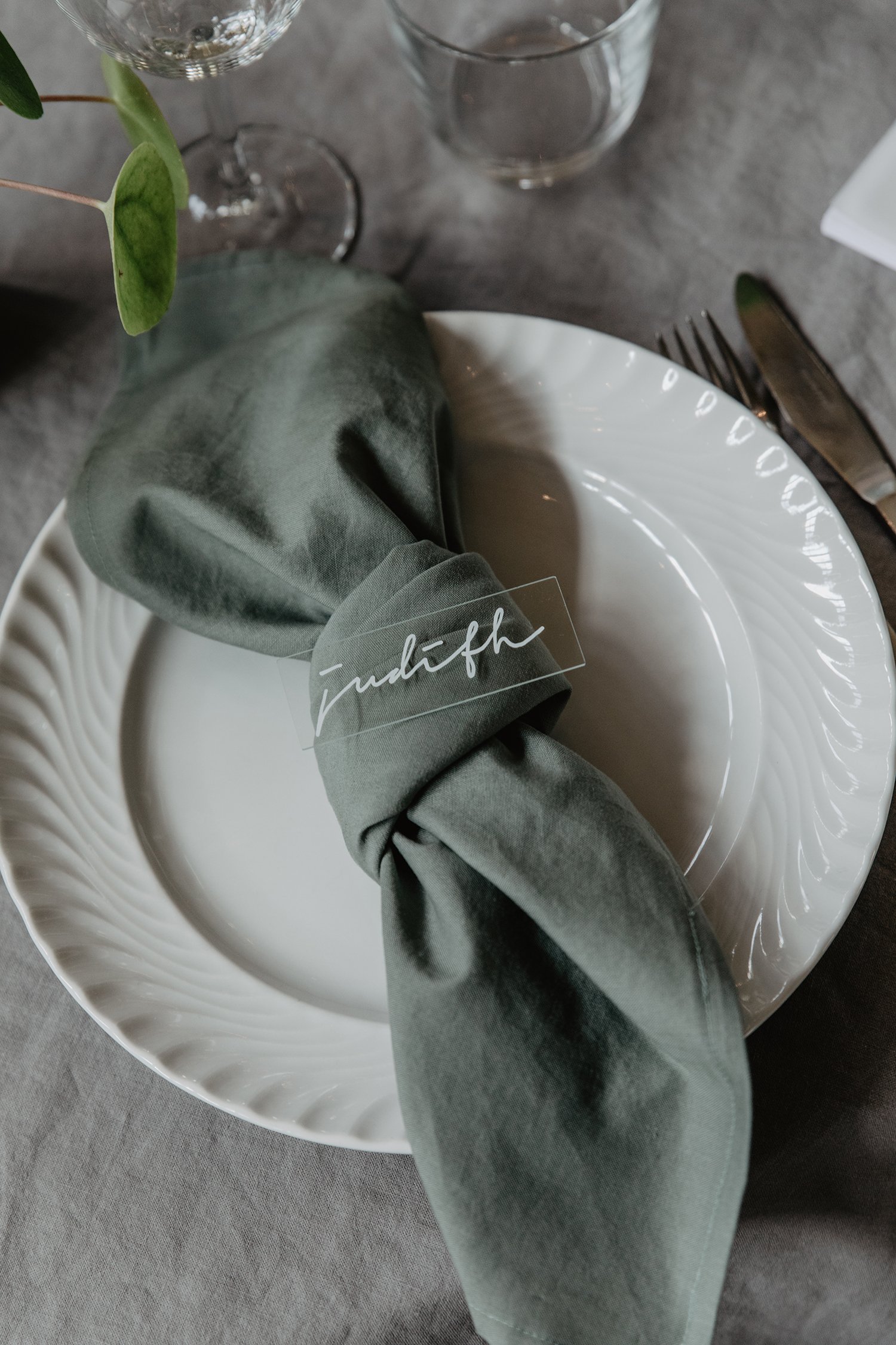 En vit tallrik med ett grönt tygservett knutet runt en platskort som har namnet 'Judith' skrivet i vitt på en bröllopsfest.