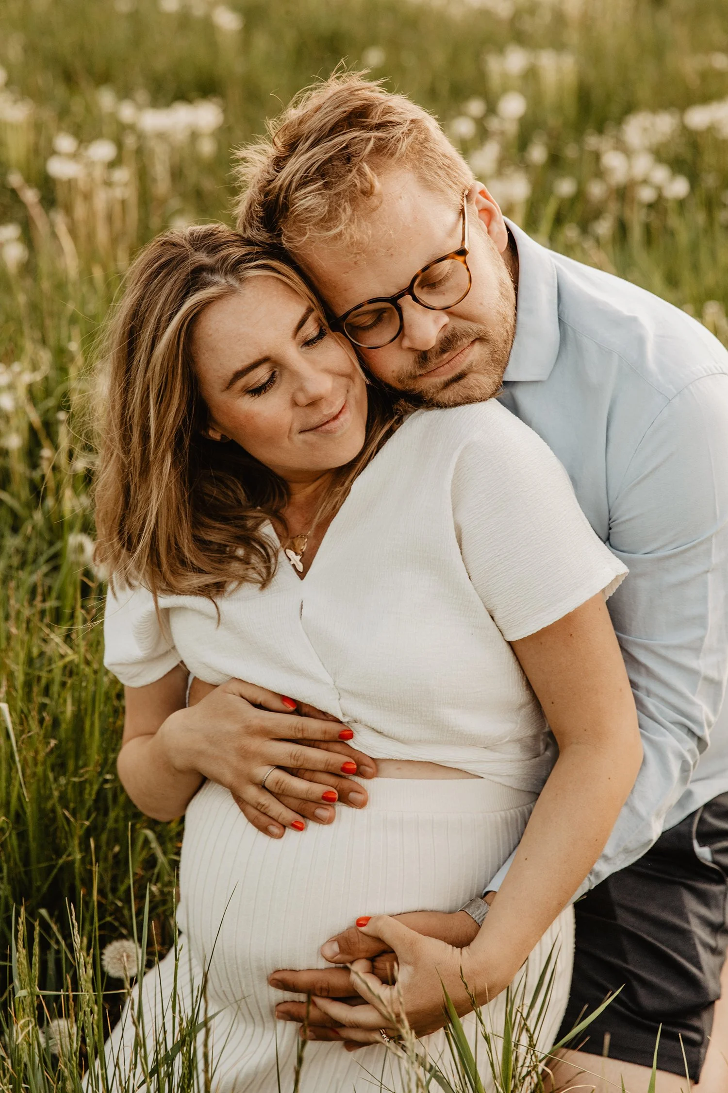 Par i en sommaräng, där kvinnan är gravid och de håller om magen, båda ler lugnt.