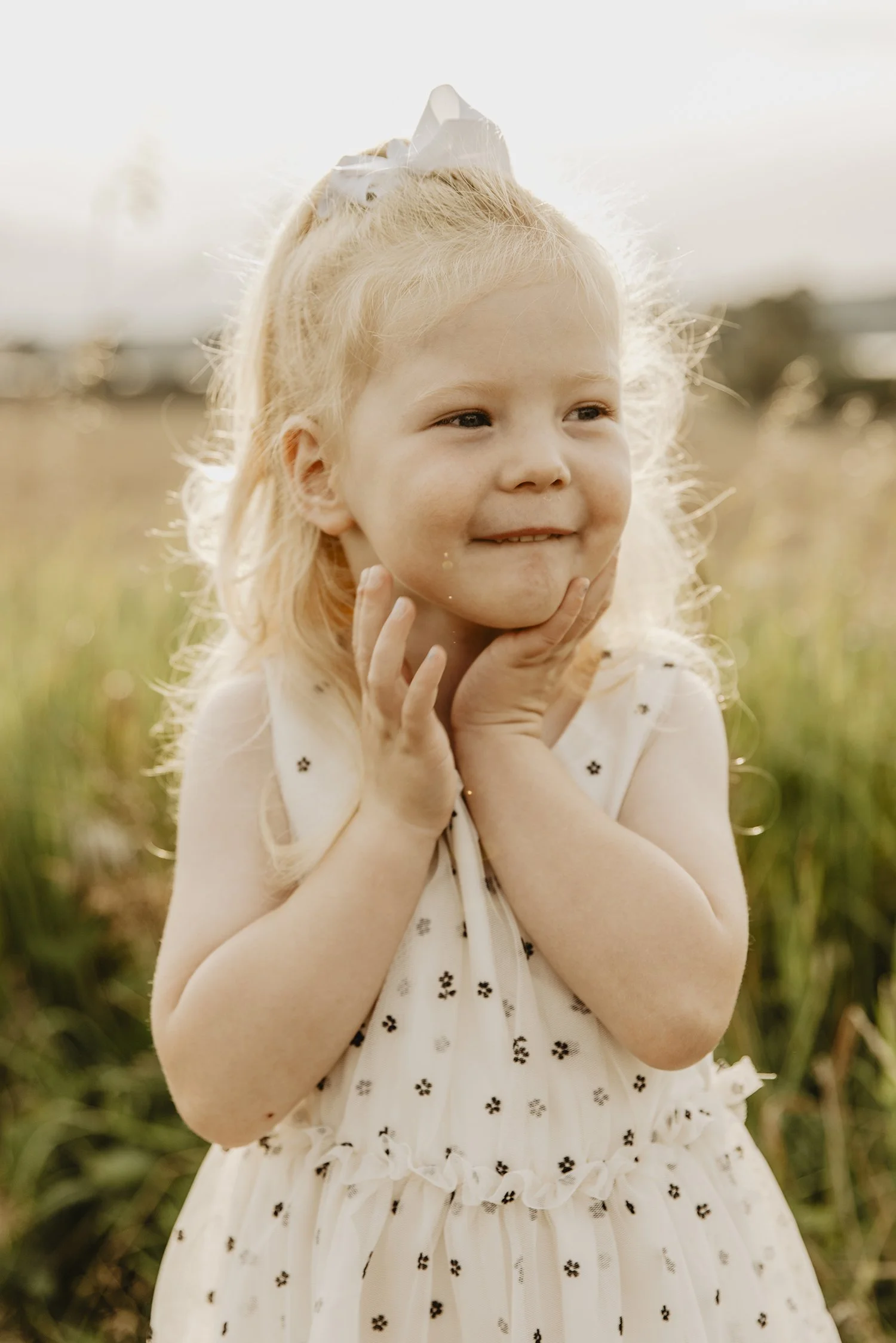 En ung flicka med blont hår som bär en vit klänning med små svarta blommor, står utomhus i ett fält i solnedgången, hon ler och håller ansiktet med händerna.