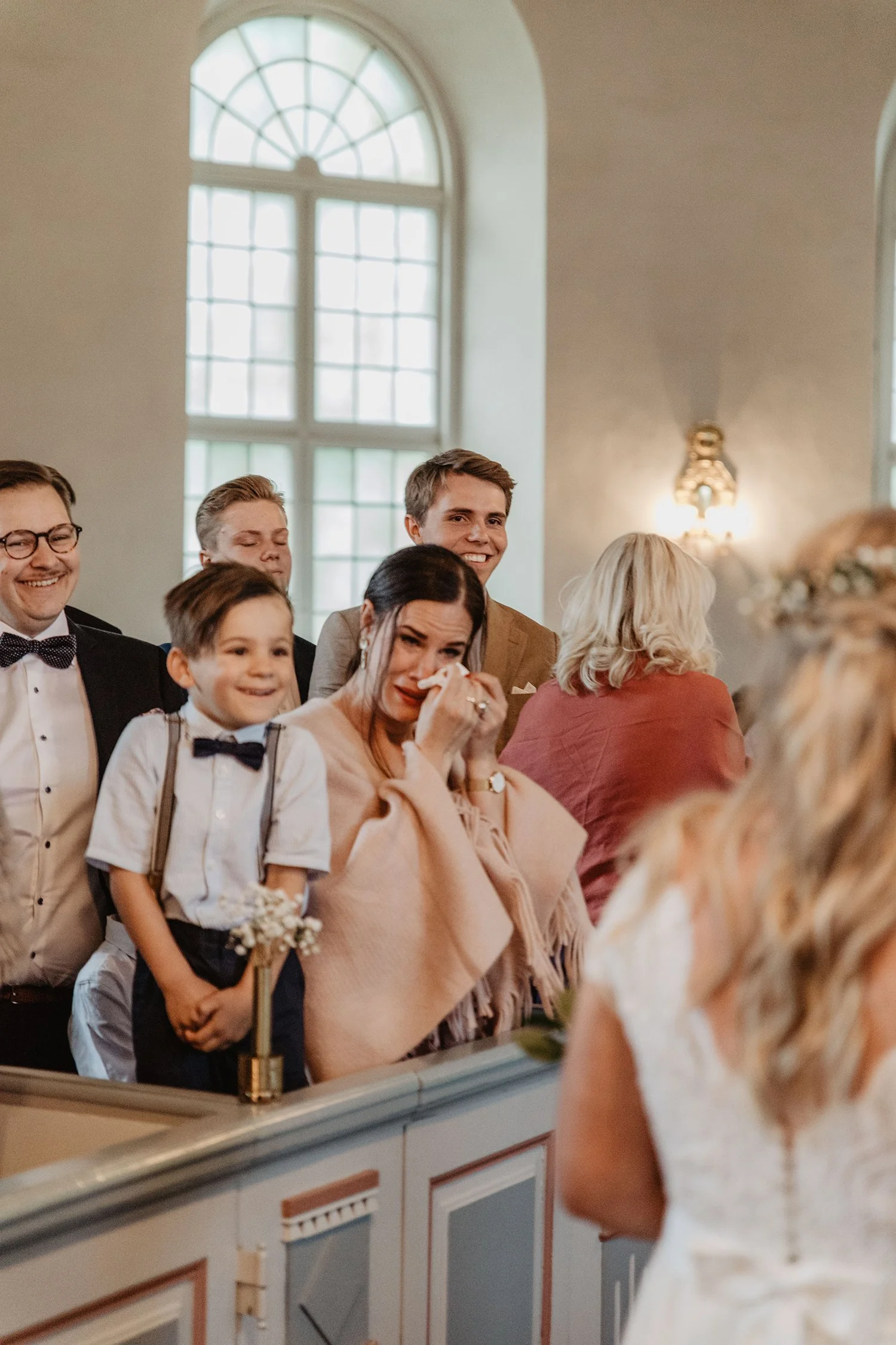 En grupp människor i kyrkan när brudparet går fram till altaret. En kvinna torkar sina tårar inför kameran på fotograferingen.
