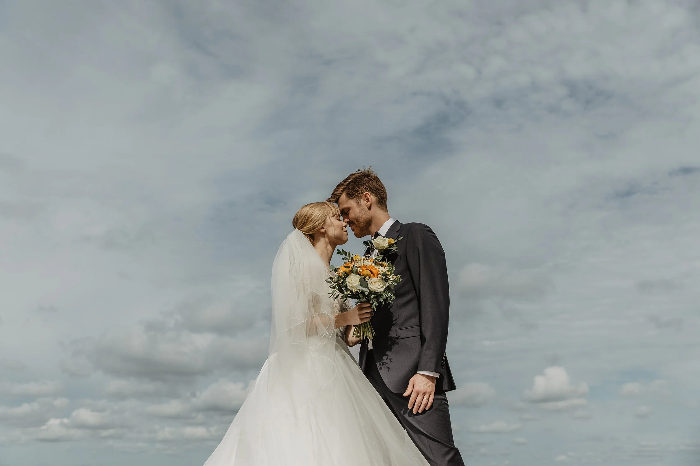 Nygift par som står nära varandra med pannan mot varandra, under en molnig himmel. Kvinnan bär en vit brudklänning och håller en bukett med vita och gula blommor, medan mannen är i en mörk kostym.
