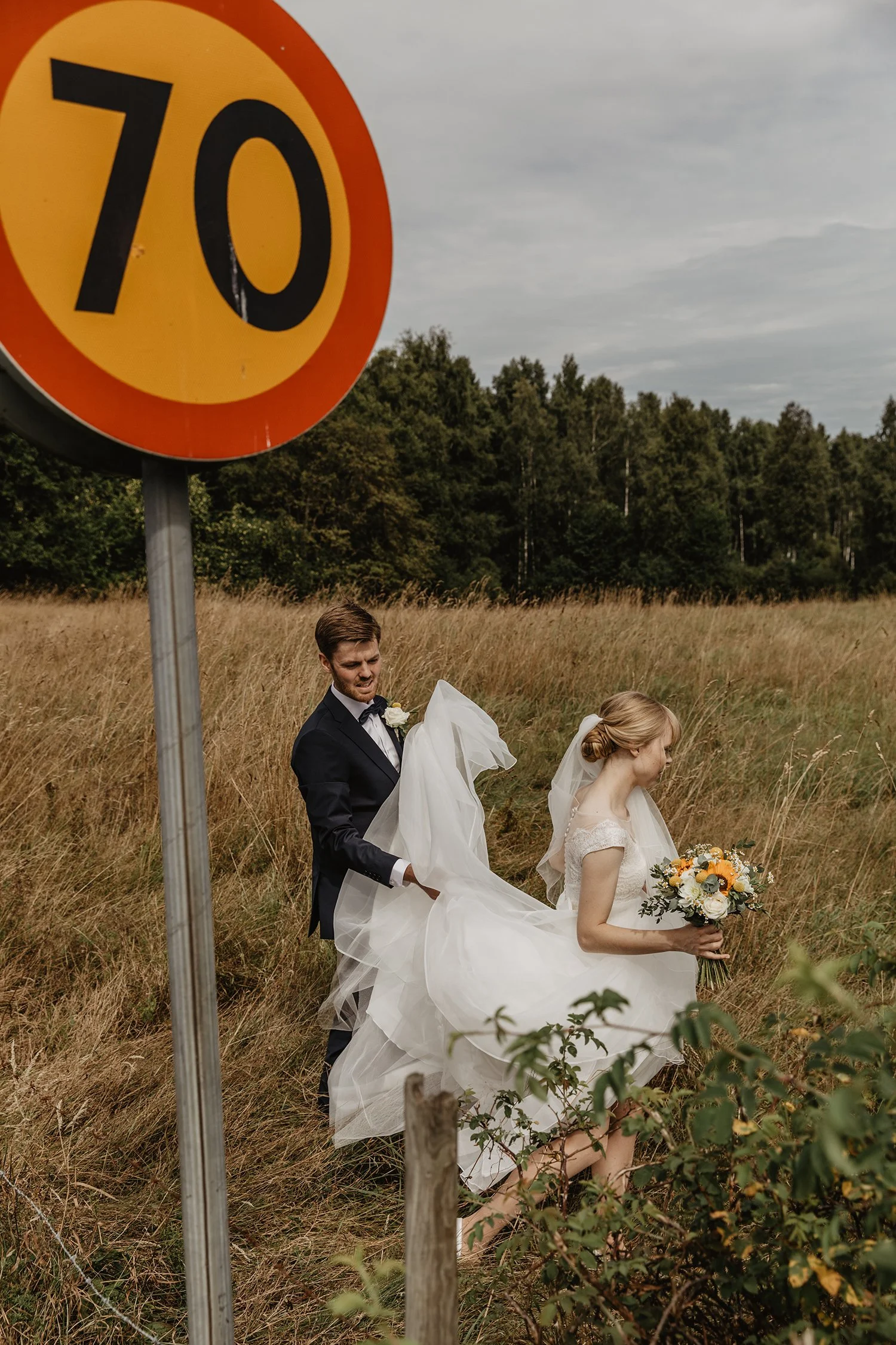 En brud och en brudgäst står i ett fält, där bruden bär en vit bröllopsklänning och vilar på brudgängets arm och håller en bukett med gula och vita blommor, medan han hjälper henne att flytta hennes klänning.