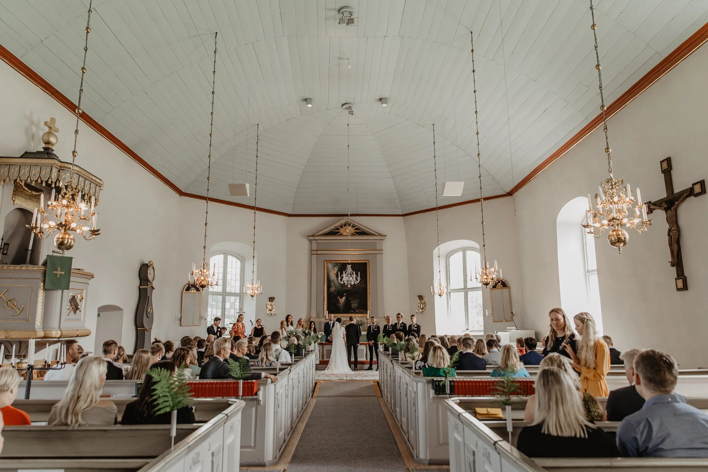 En vigsel i en ljus kyrka med gäster, bruparet står med ryggen vända mot kameran längst fram vid altaret. BIlden är tagen under en bröllopsfotografering.