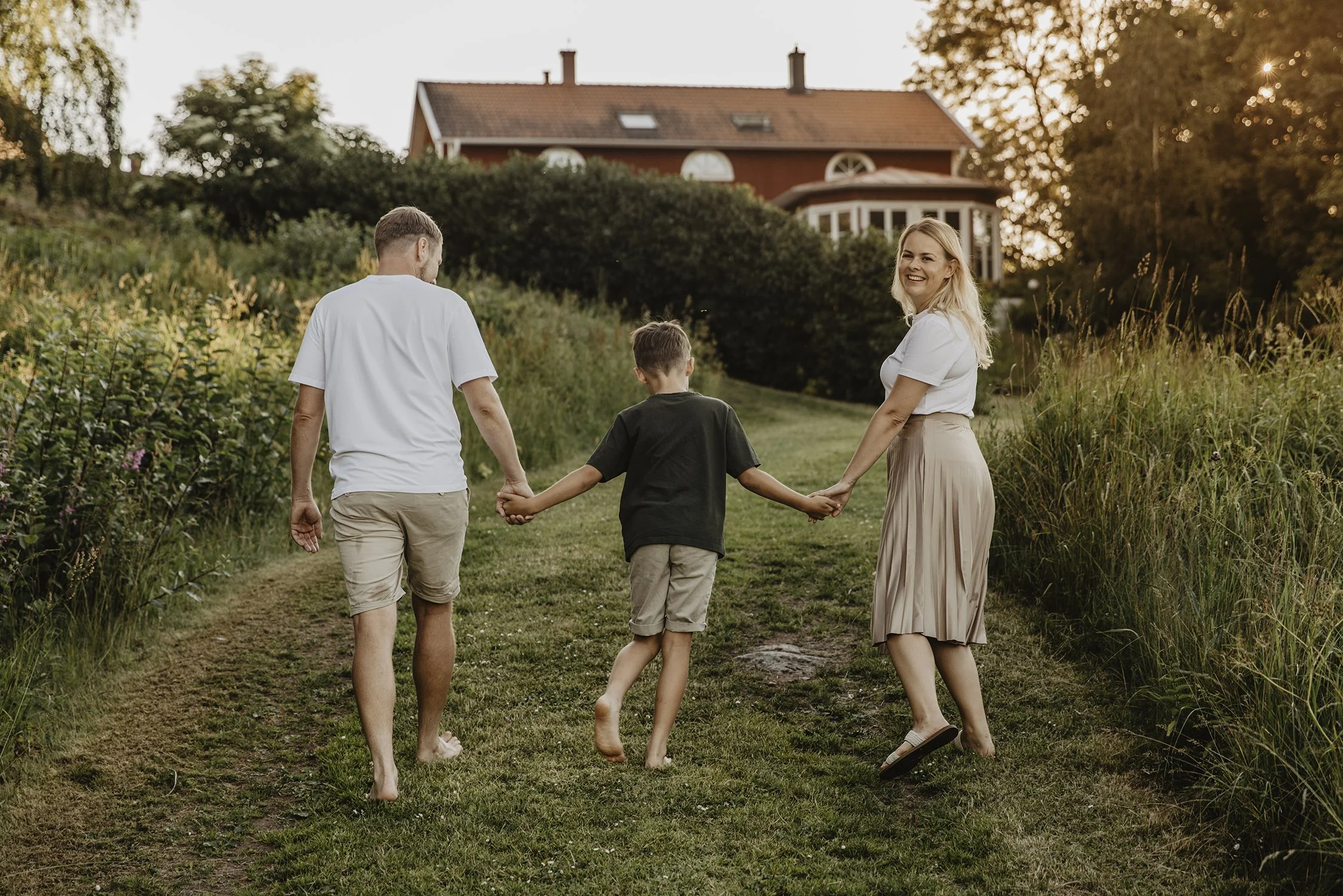 Familj som går hand i hand på en gräsbevuxen stig utanför Gränna vid Bun i solnedgången, med ett hus i bakgrunden.