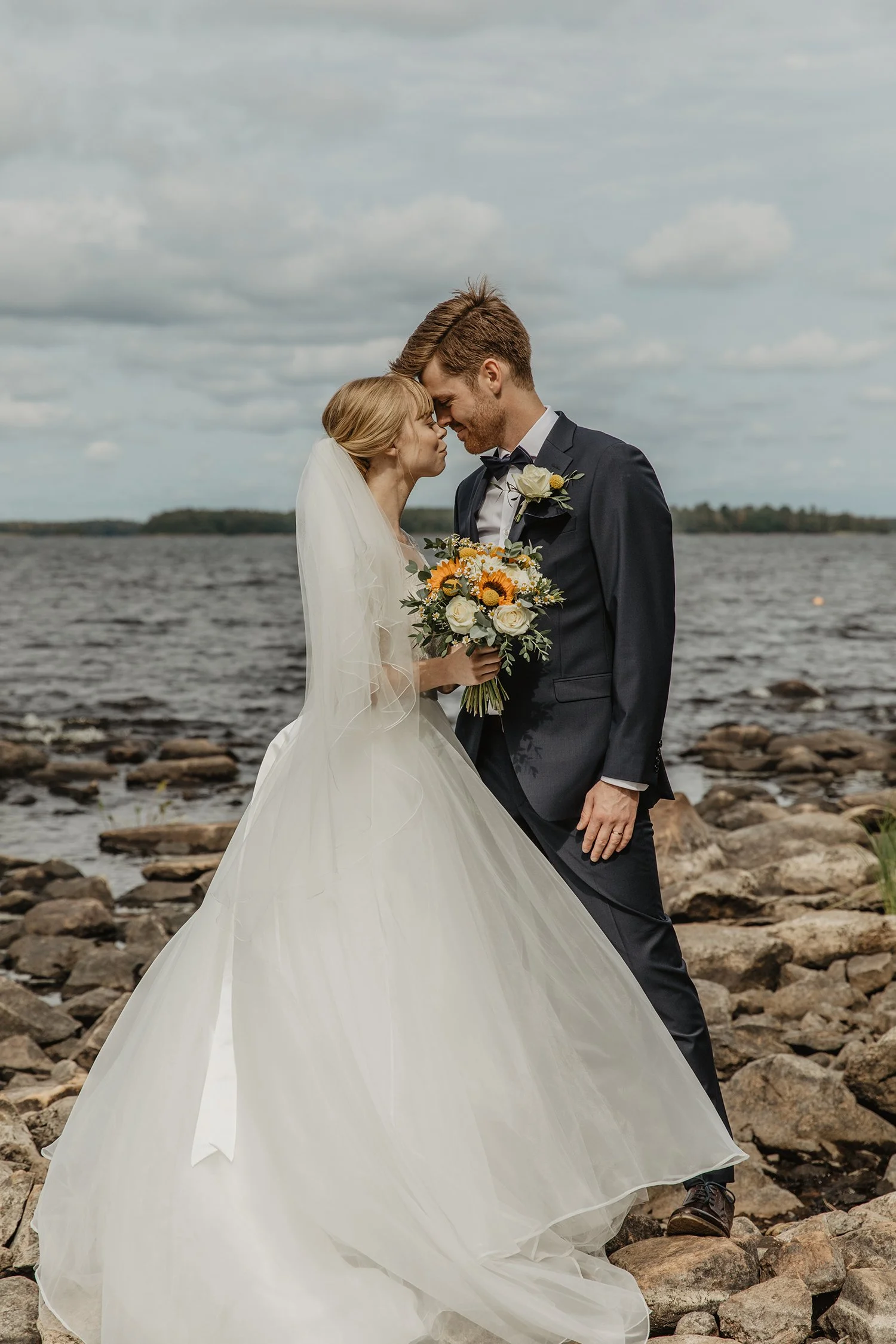Brudpar på stranden vid vattnet, leende och nära varandra, de håller en bukett med solrosor och vita blommor.