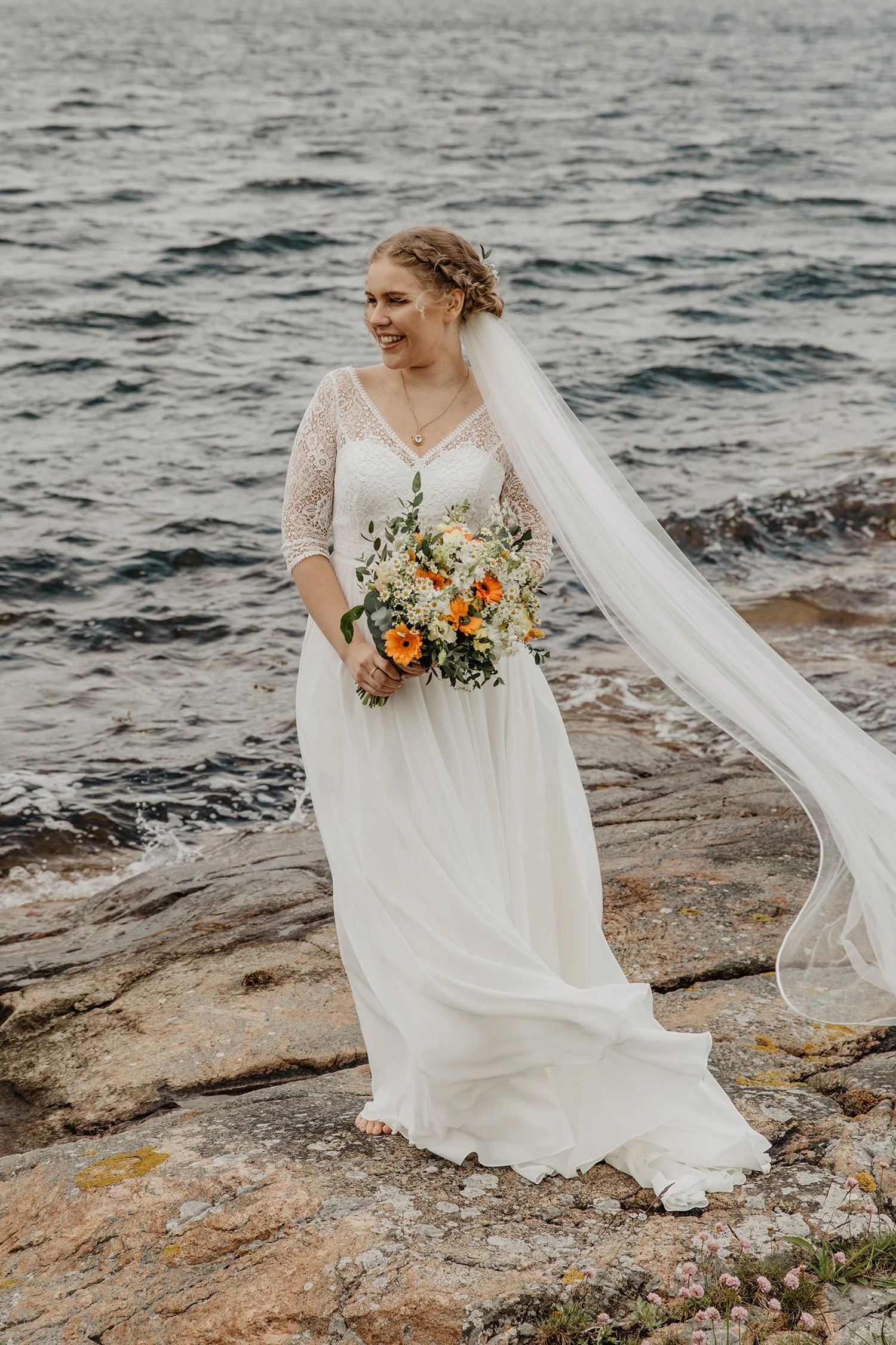 En brud i vit bröllopsklänning står på en klippa vid vattnet, håller en blomsterbukett och ler.