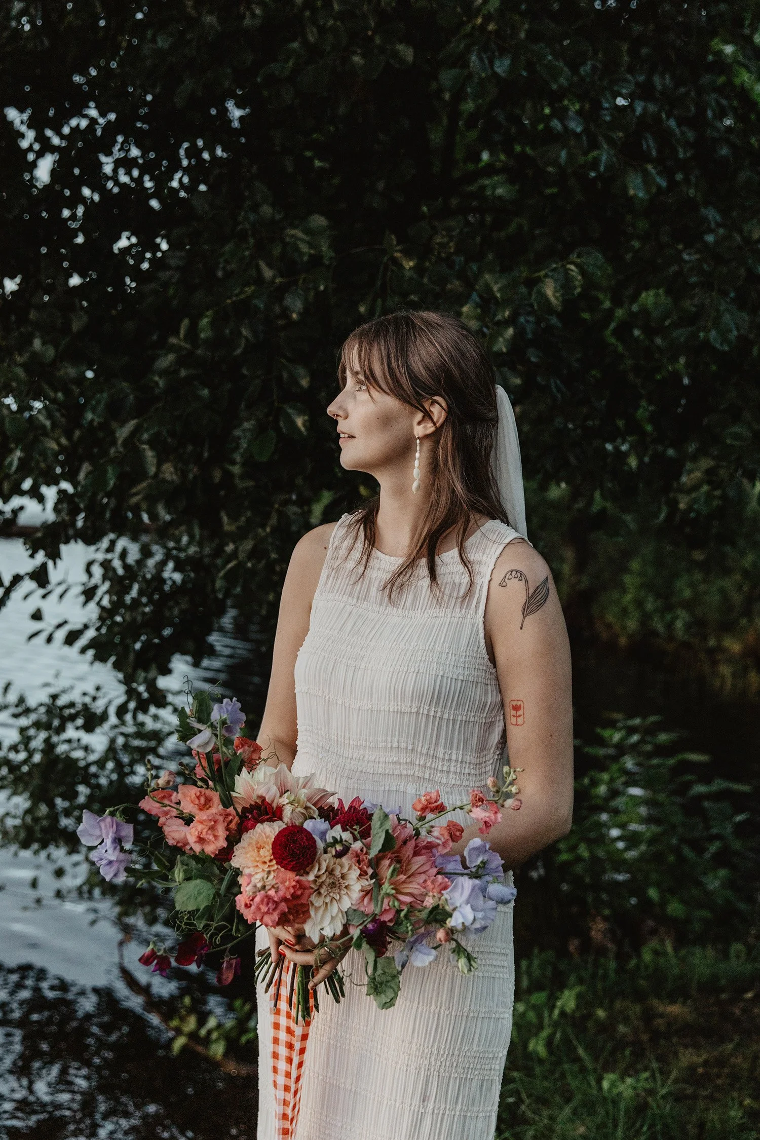 En kvinna i vit klänning håller en bunt färgglada blommor vid vatten omgiven av gröna träd.