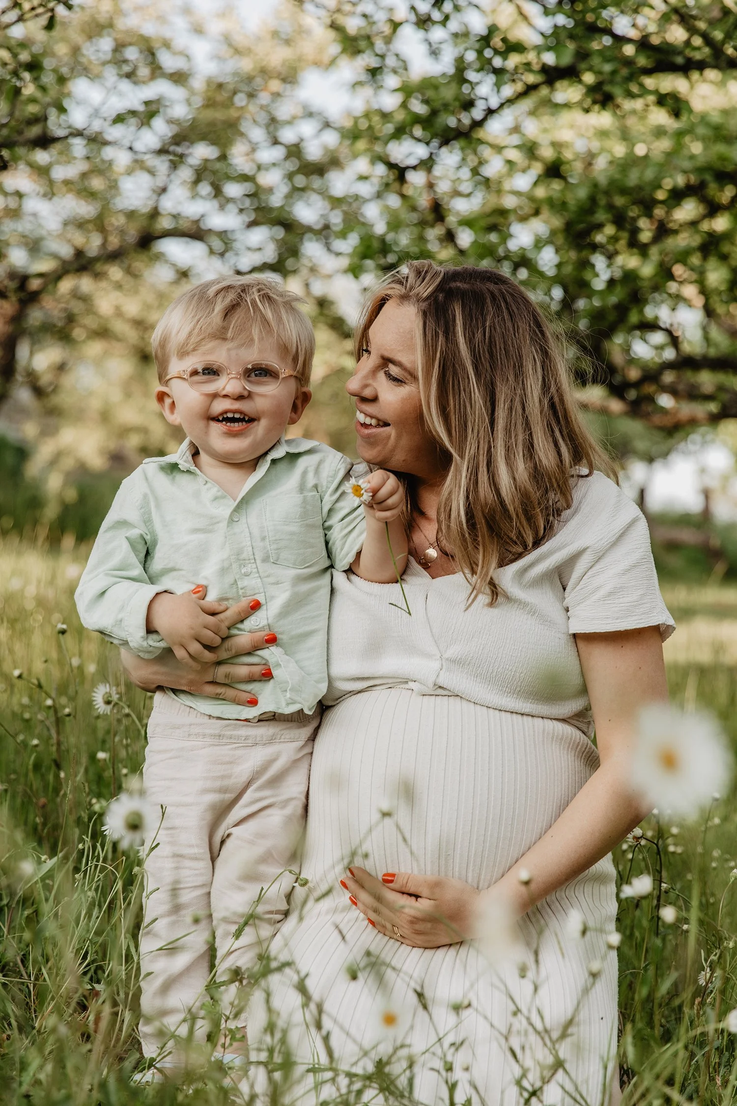 En gravid kvinna i en blomsteräng med ett barn i hennes fång, som ler och har glasögon, under ett träd.