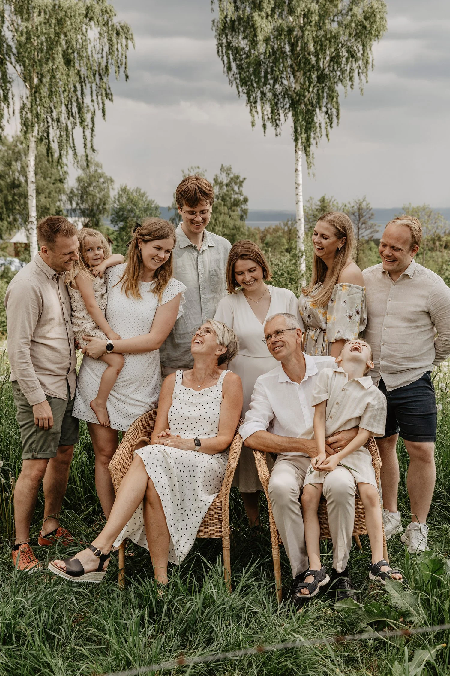 En grupp människor utomhus, alla skrattar och ler medan de samlas runt två personer som sitter i stolar i mitten. Det är en grön och skogig miljö med träd och gräs.