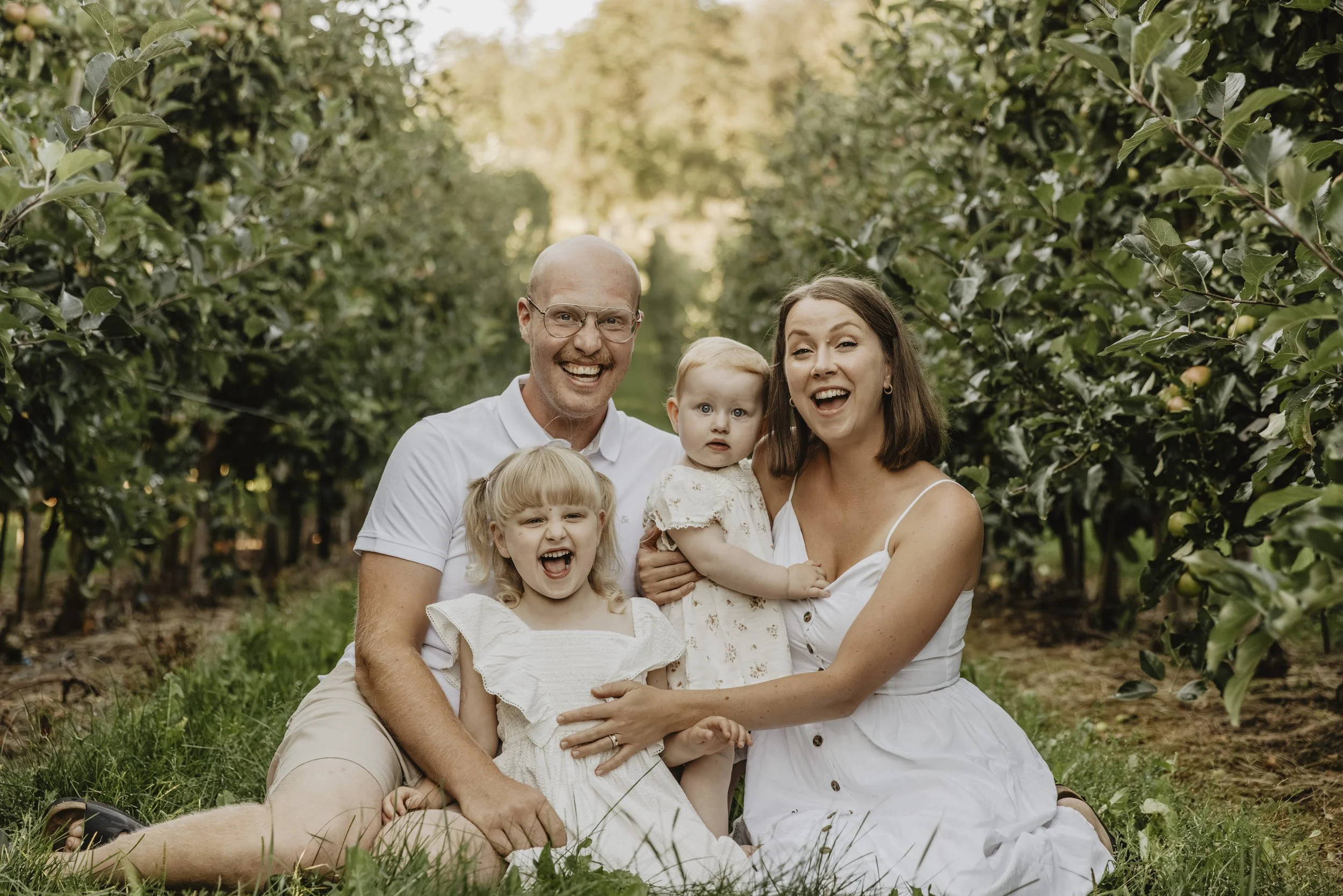 En glad familj med tre barn, sittandes på gräset i ett äppelträdgård.