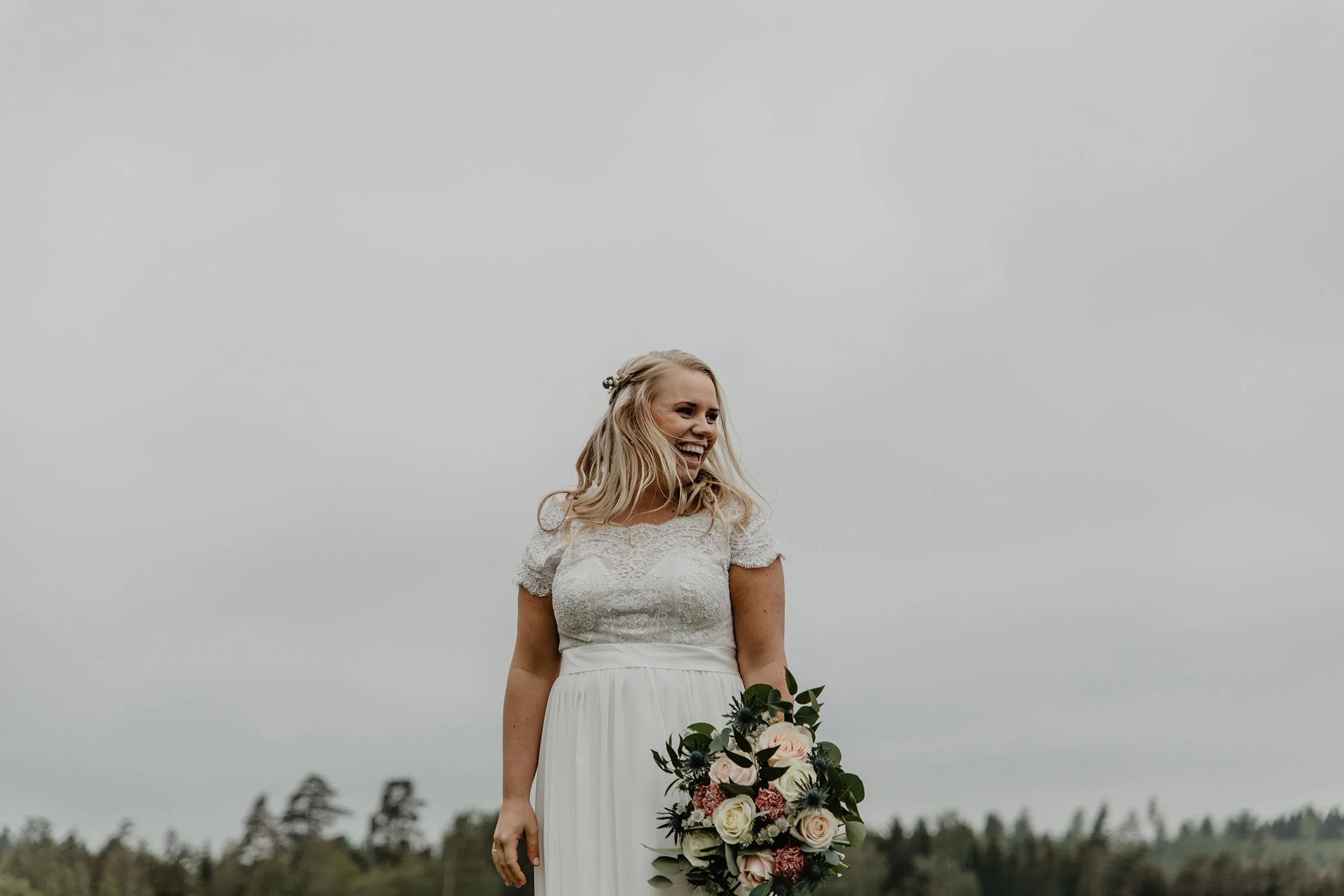 En glad brud står utomhus med ett bukett med vita och rosa blommor, mot en grå himmel och ett landskap av träd under en bröllopsfotografering.