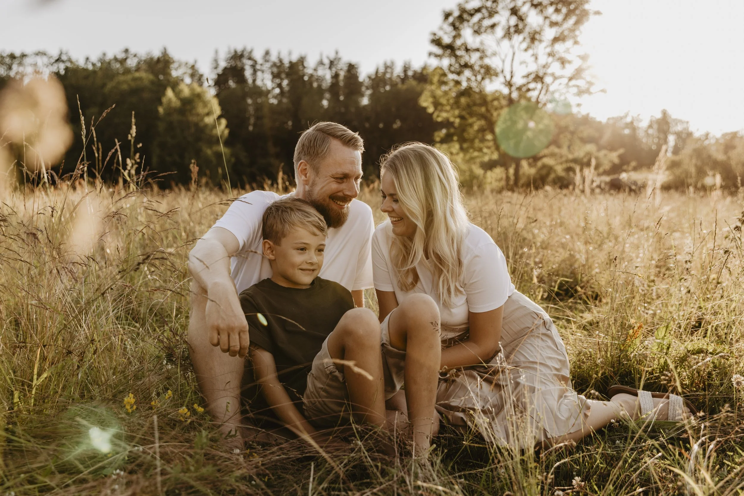 En familjefotografering i Gränna med två föräldrar och ett barn. De sitter i en hage med högt gräs i solnedgången och är glada.