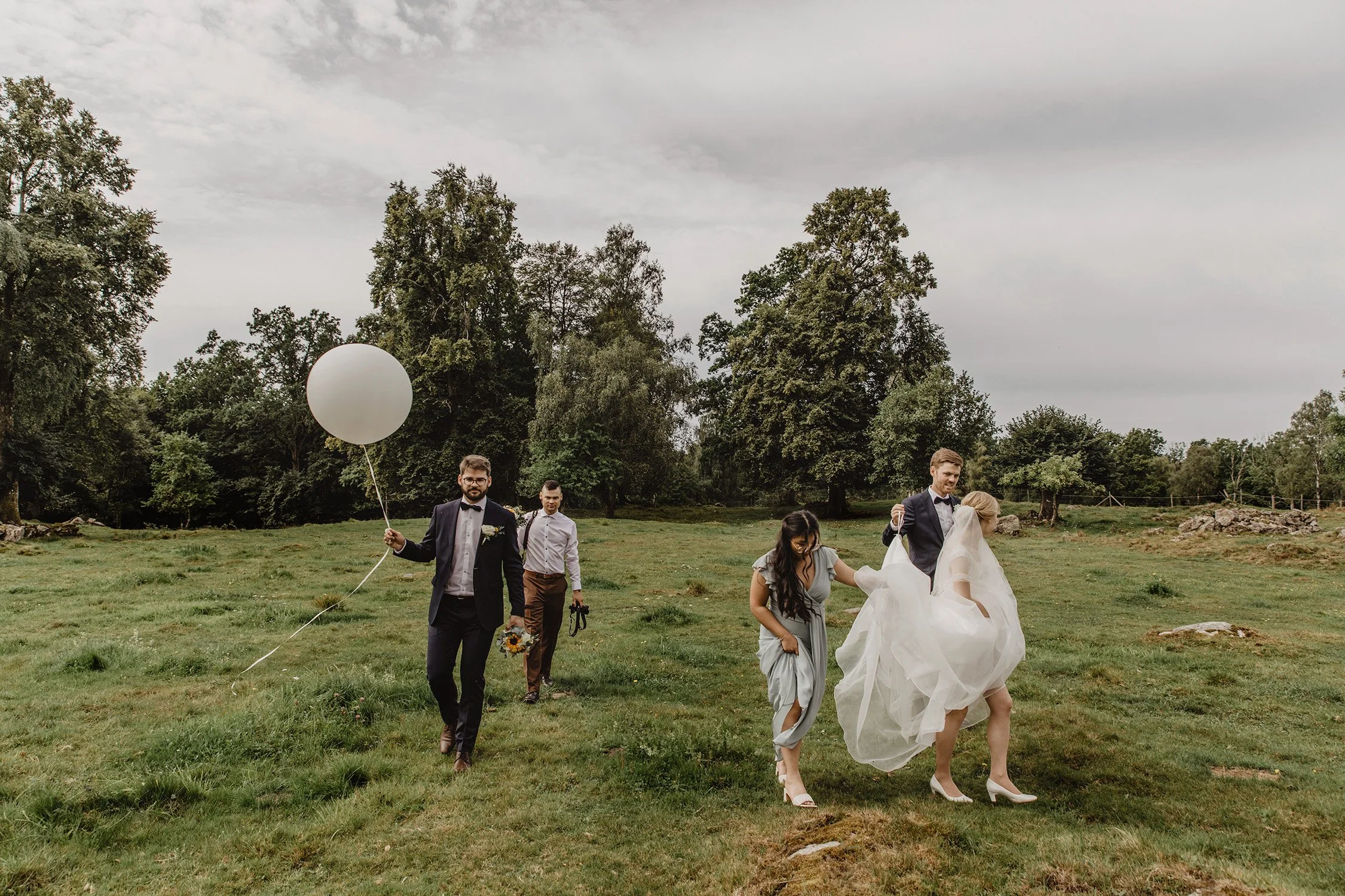 En grupp människor, inklusive en brud och en groom, är ute på en grön ängen med träd i bakgrunden. Bruden bär en vitt brudklänning och högklackade skor, och groomen bär en svart kostym. Någon håller i en stor vit ballong.
