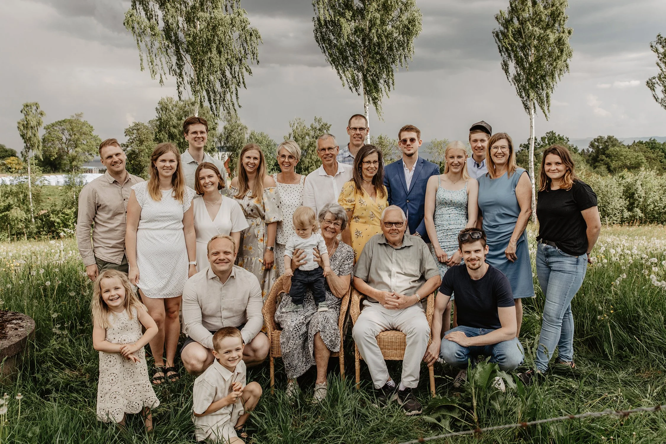En stor grupp människor samlade för en familjefoton i en grön, blommande utomhusmiljö med träd och en övercast himmel i bakgrunden.