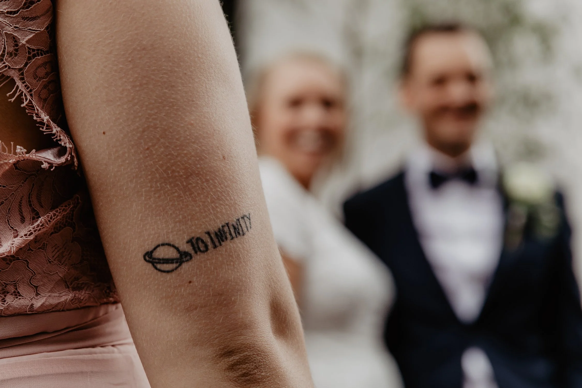 En person med en tatuering av en liten planet och texten "TO INFINITY" på armen. I bakgrunden syns ett suddigt brudpar som ler.