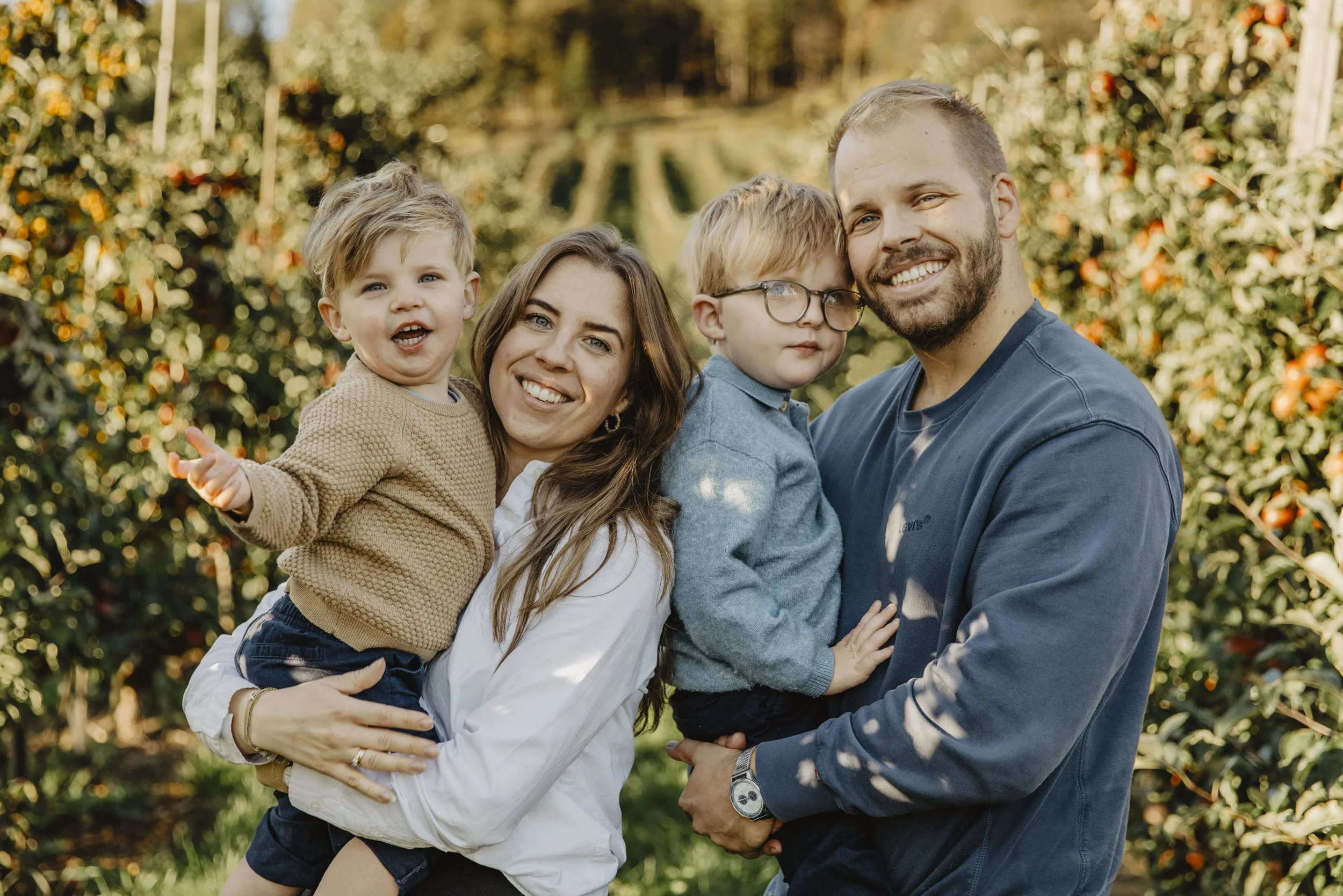 Familj av två vuxna och två barn utomhus i ett äppelträdgård på hösten, ler och poserar för foto.