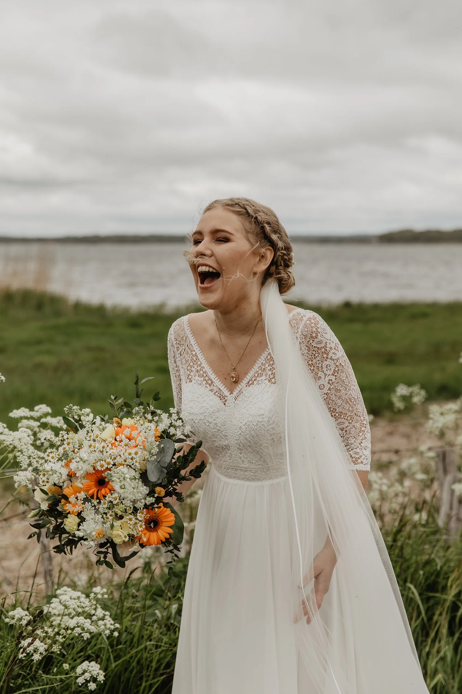 En brud ler på en utomhusplats med vattendrag i bakgrunden, bär en vit brudklänning med spetsärmar och håller en blomsterbukett i orange och vitt.