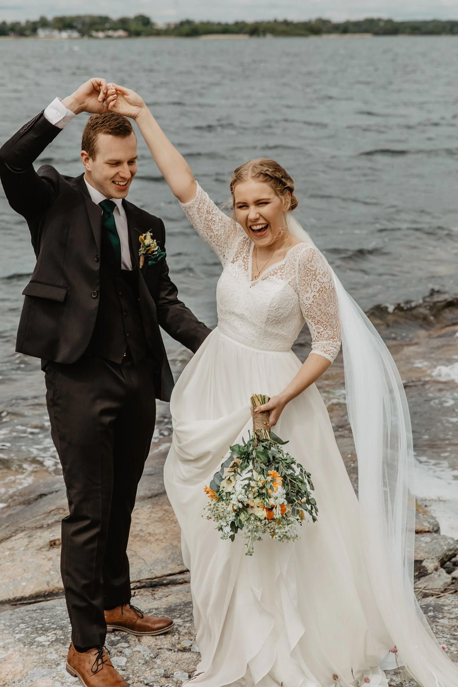 Nygifta par på stranden, mannen håller igenom tjejens hand och hon håller en bukett blommor.