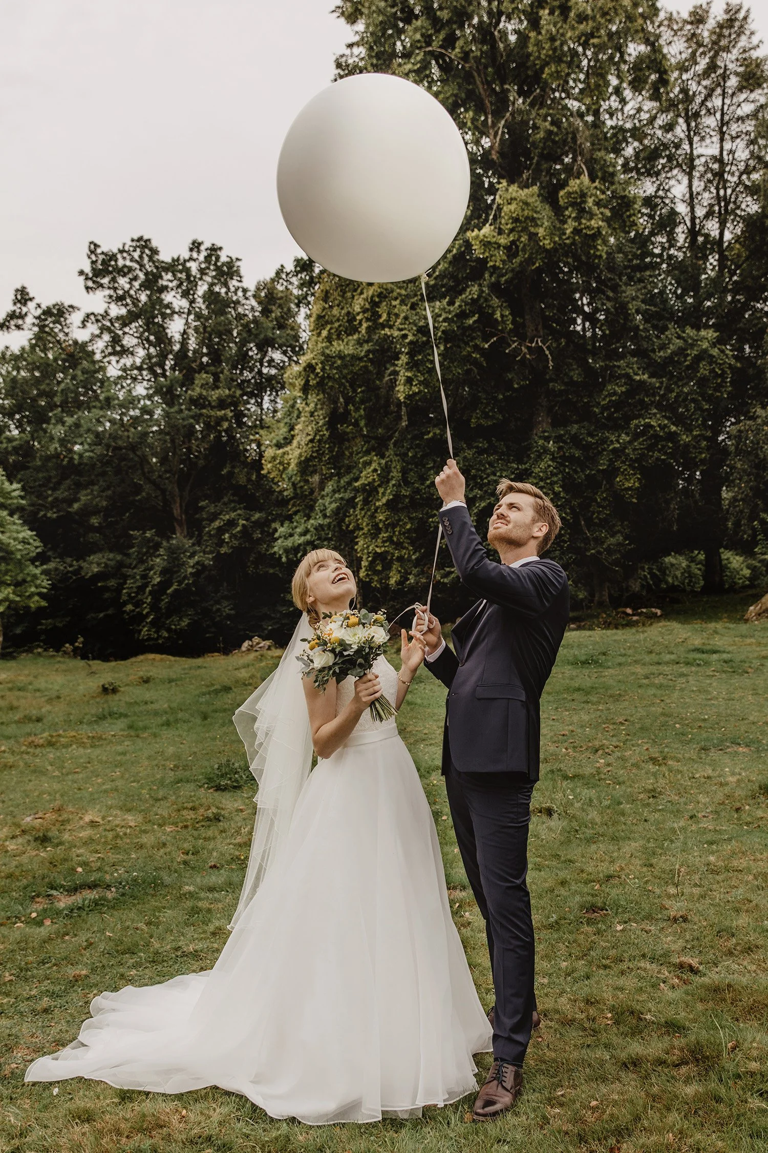 En brud och en brudgum utomhus, där brudgummen släpper en vit helium-helikopterballong, medan bruden håller en blombukett och tittar på ballongen.
