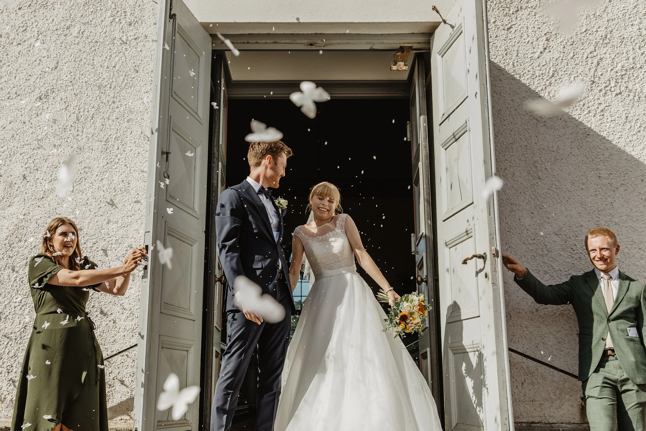 Nygift par står i kyrkporten omgiven av sina gäster som kastar ris. Bruden ler glatt och håller en blomsterbukett, medan brudgummen ser på henne.