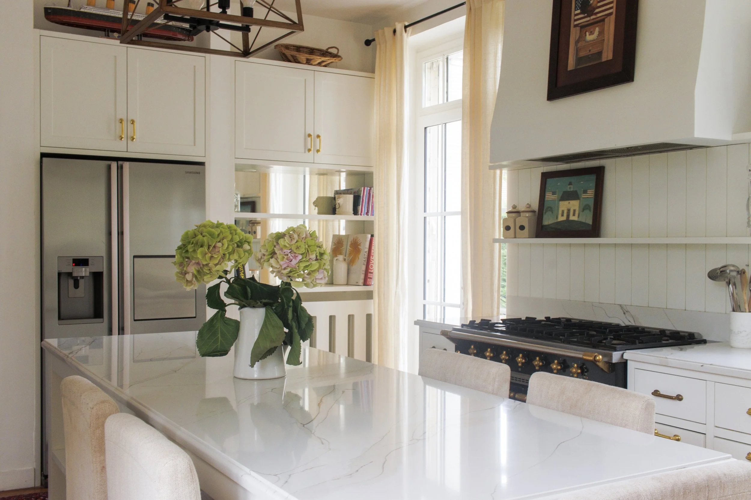 A modern kitchen with white cabinets, a stainless steel refrigerator, a white marble island with a vase of green and pink hydrangeas, a large window with cream curtains, and framed artwork on the wall.