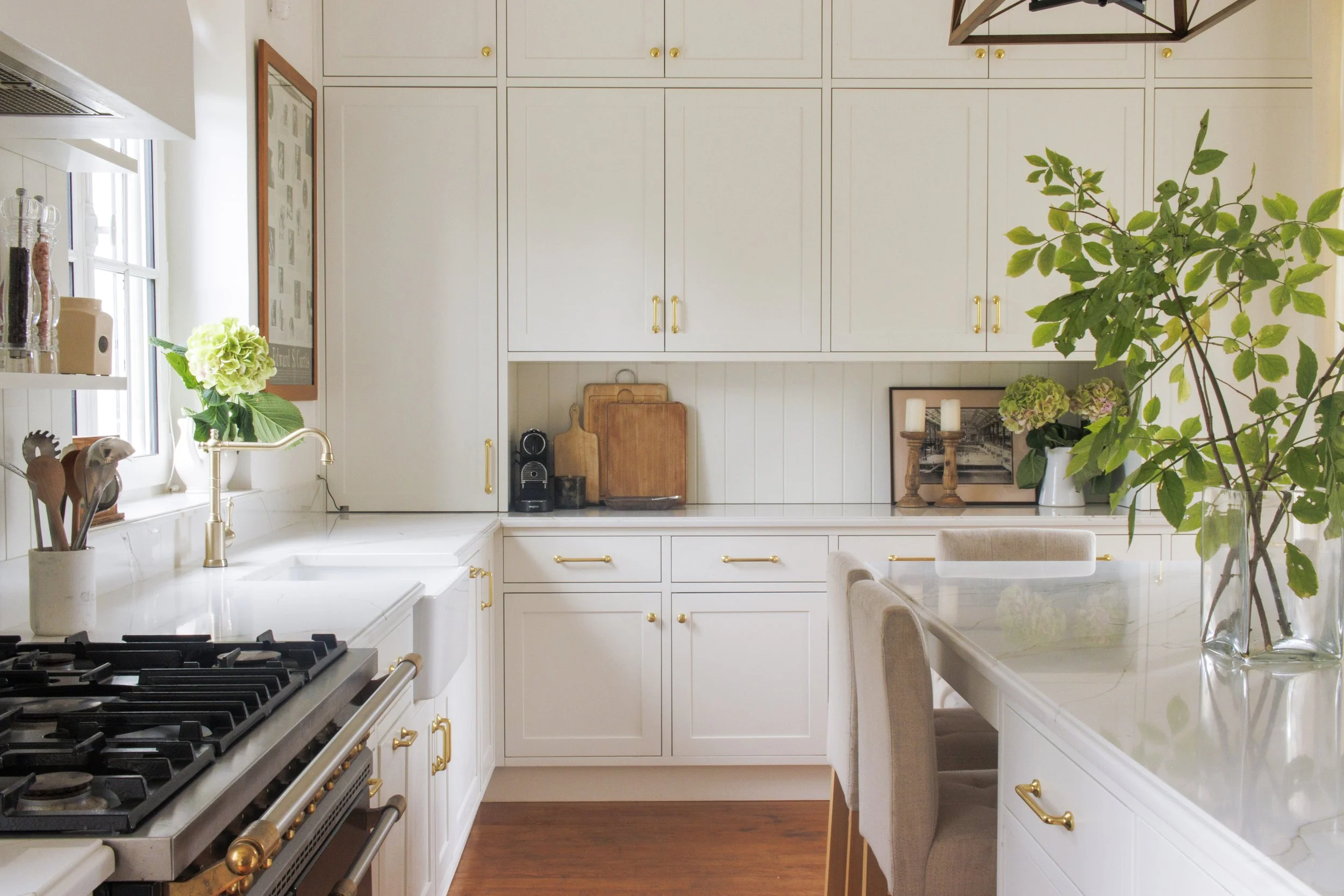 Bright white kitchen with white cabinets, gold hardware, a gas stove, a farmhouse sink near a window with a plant, and a marble countertop island with beige chairs. Decorative items include cutting boards, candles, a framed photo, and vases with gree