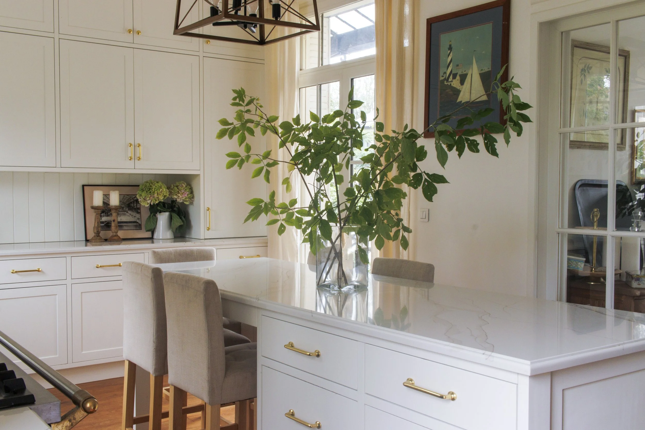 Bright kitchen with white cabinetry, a marble countertop island, beige chairs, large window with cream curtains, a glass vase with green branches, and wall art of a lighthouse and sailboats.