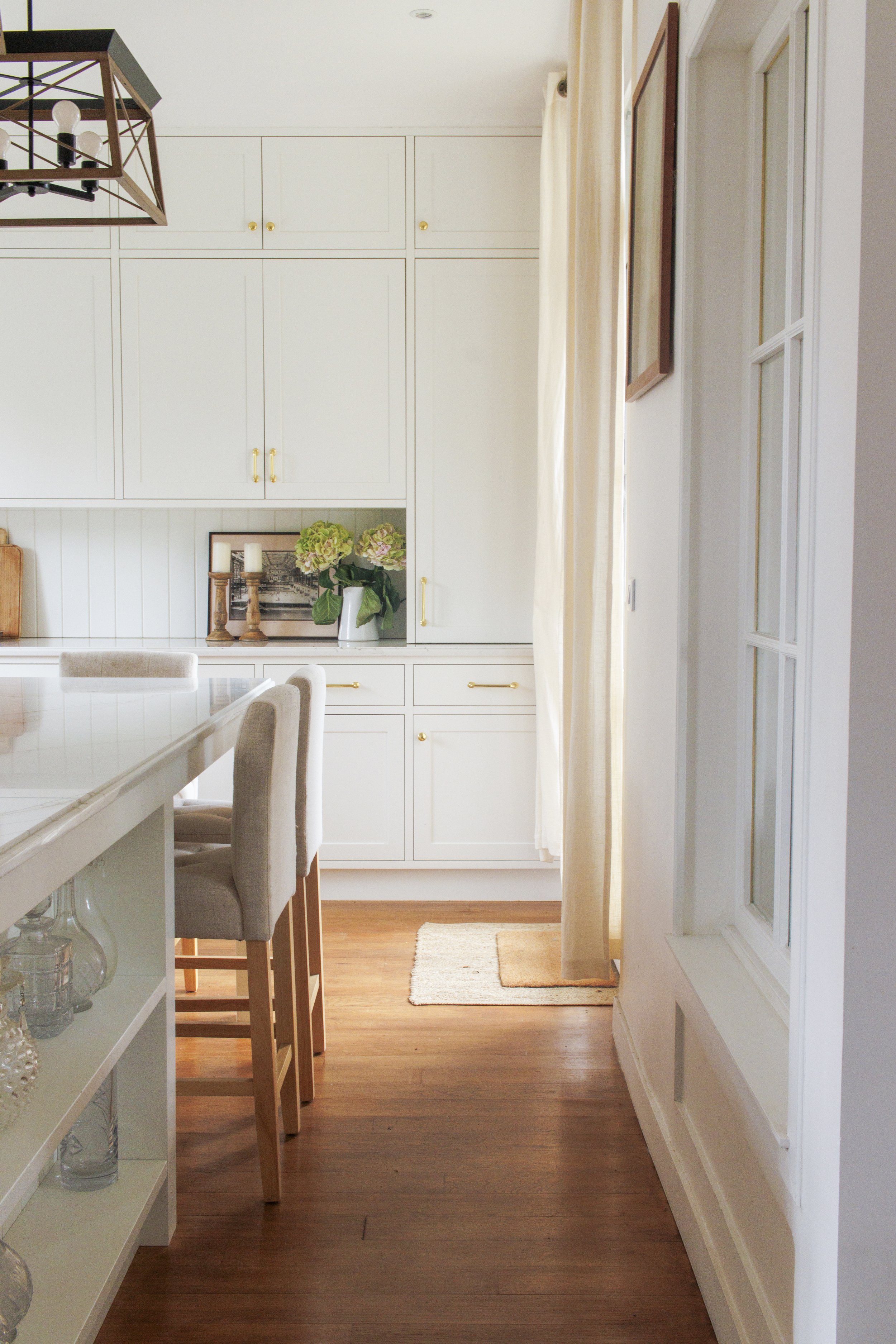 A bright, cozy kitchen nook with white cabinets, a wooden floor, light-colored curtains, and a small rug.