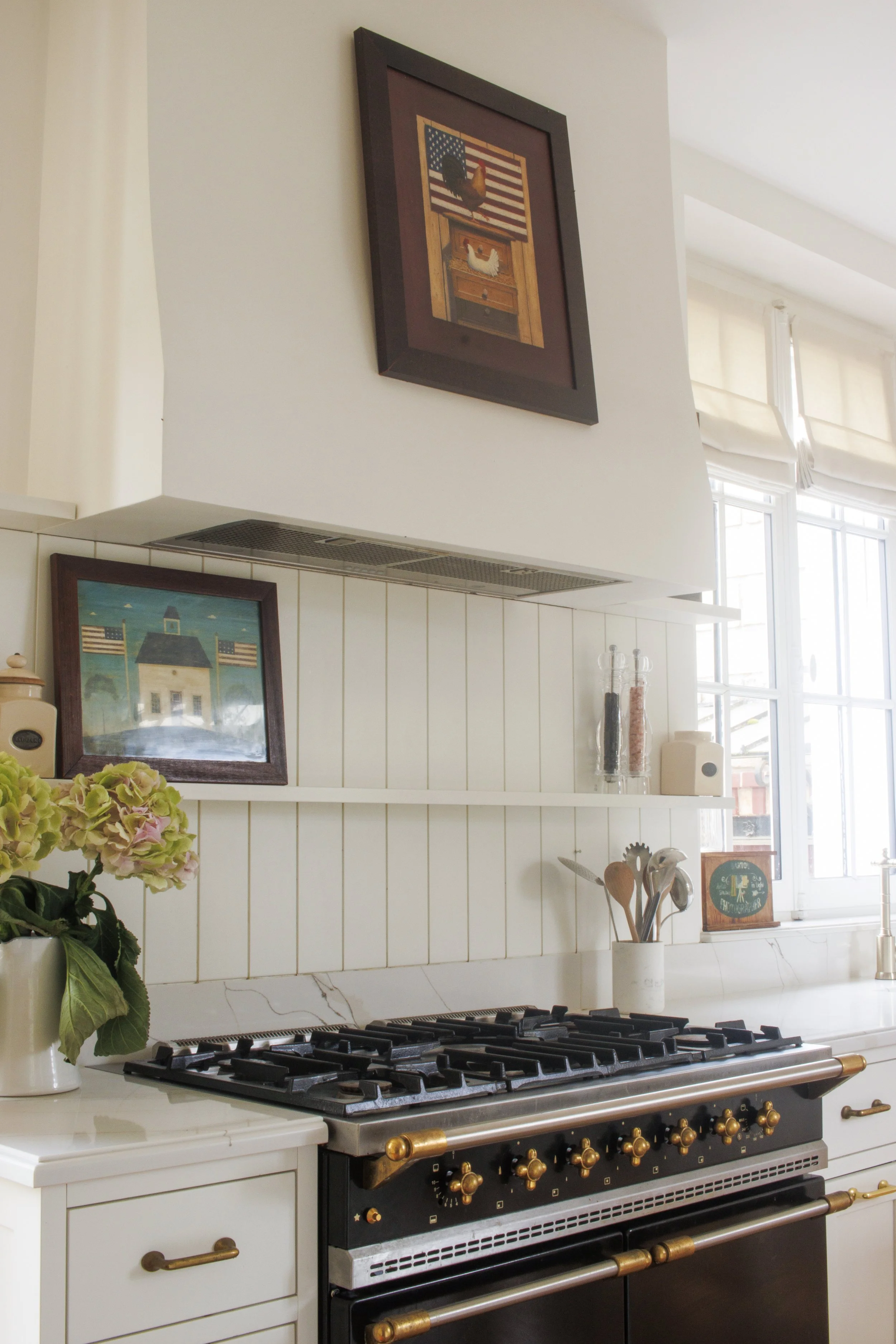 Kitchen with a black stove, a white countertop, and framed pictures, including one of a rooster with an American flag background. There are flowers, utensils, and spices on the counter near a window.