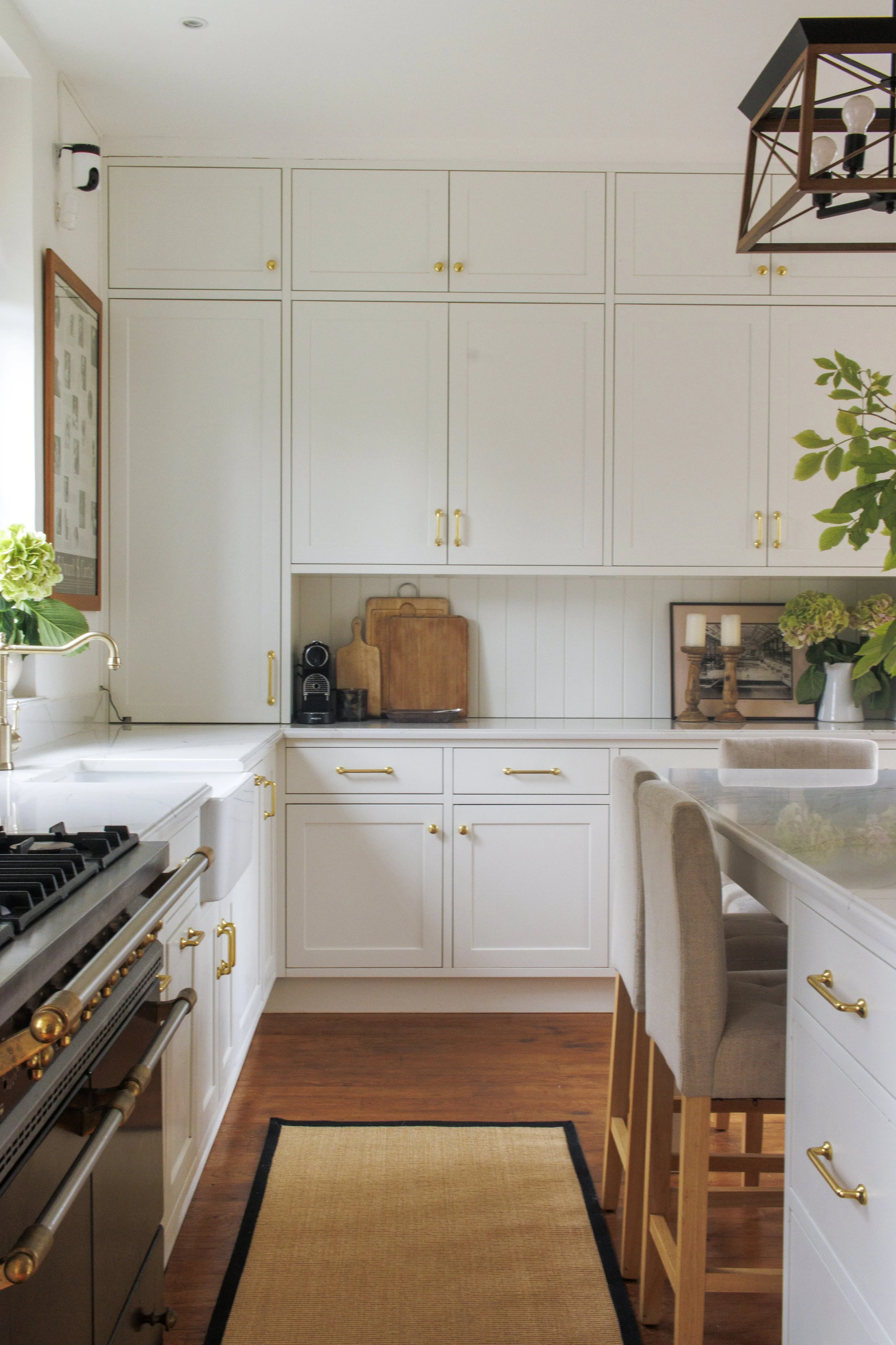 Bright, white kitchen with gold hardware, wooden flooring, and a black rug. Includes built-in cabinets, a stove, a kitchen sink, and counter space with decor and plants.