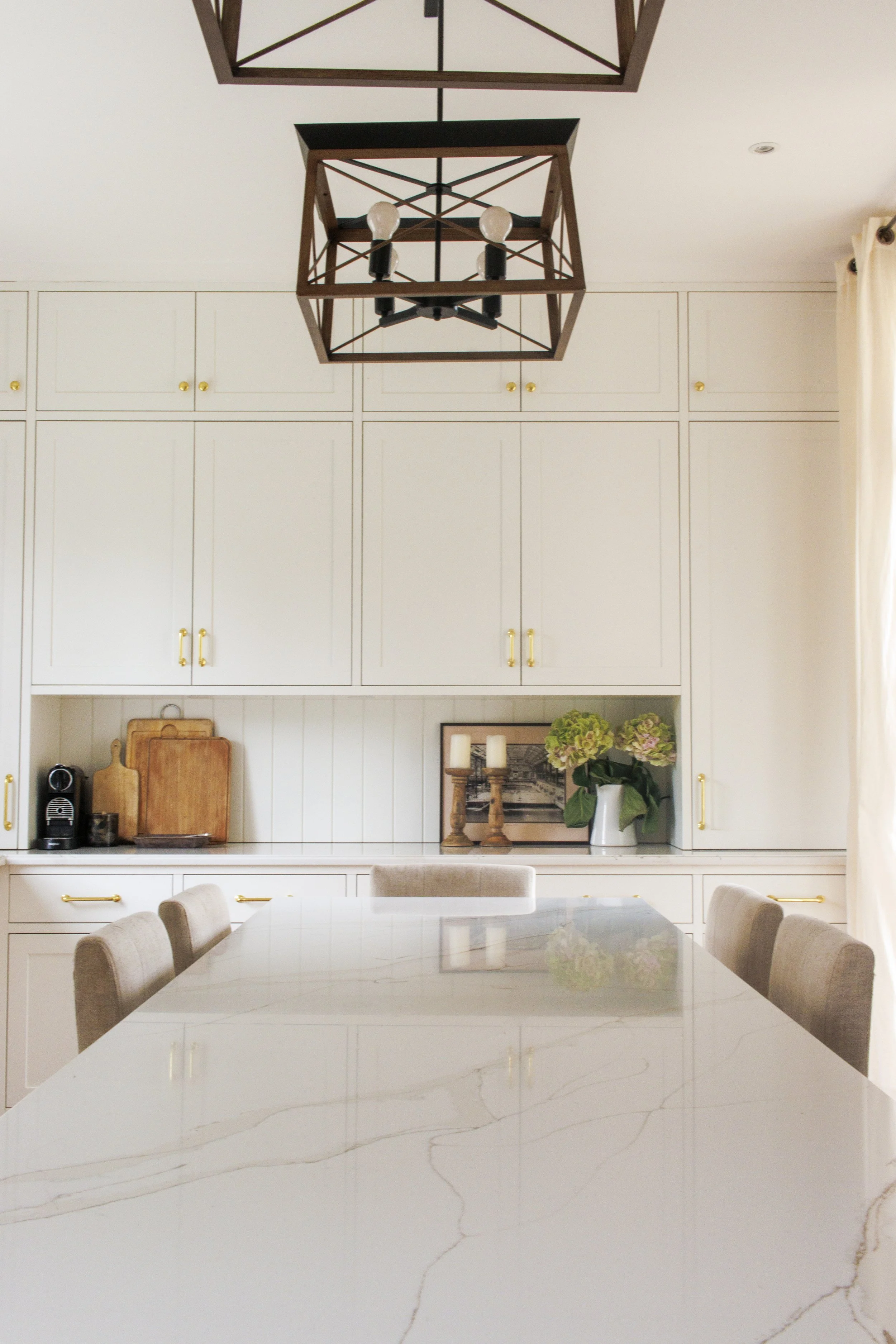 A modern dining room with a large marble table, beige upholstered chairs, white cabinets with gold handles, and decorative items like candles, framed pictures, and flowers. Modern black geometric chandeliers hang from the ceiling.