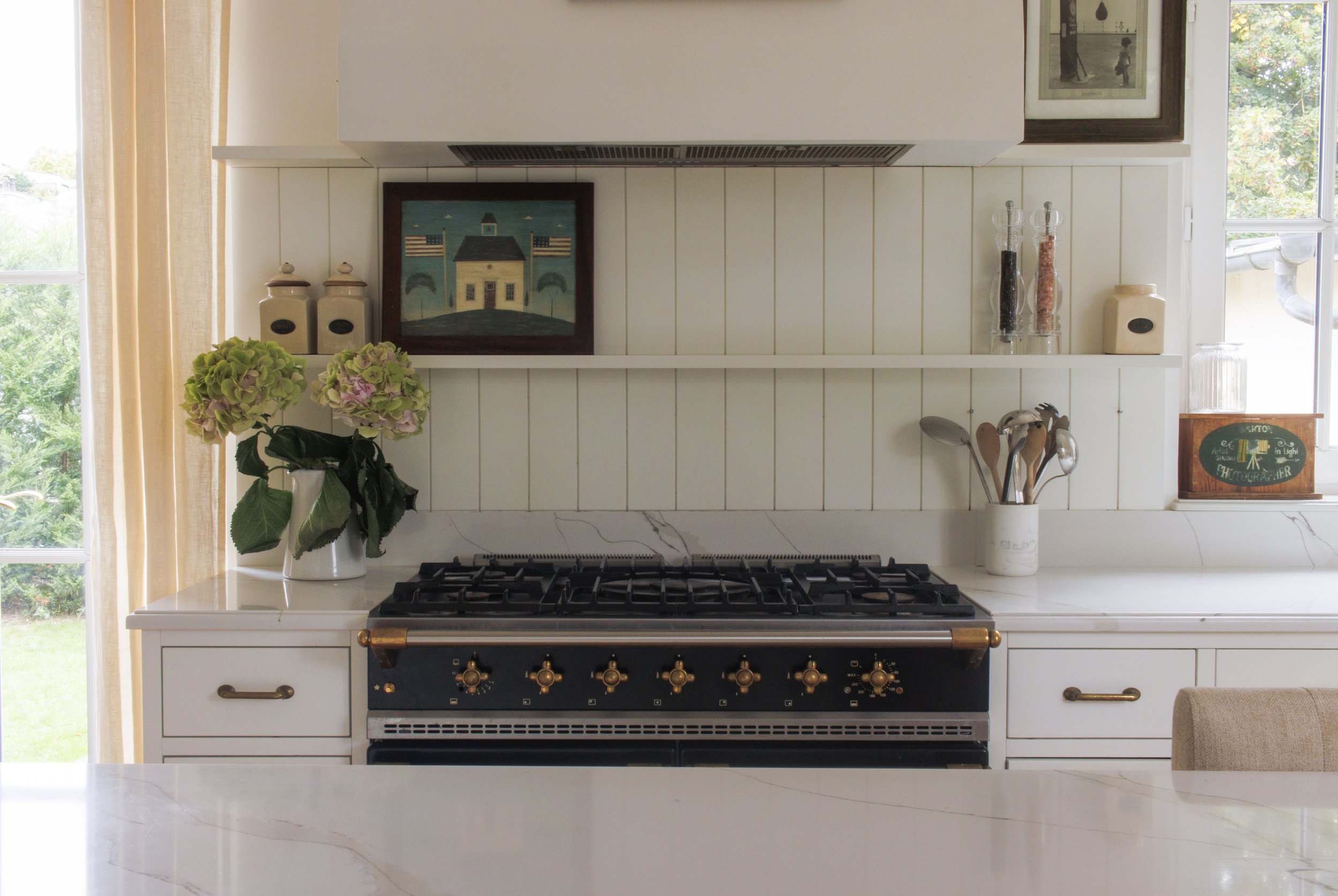 A kitchen with a white marble countertop, a white stove with gold knobs, a potted plant with pink and green hydrangea flowers, and open shelves with decorative jars, framed art, and salt and pepper shakers. There is a window with curtains on the left