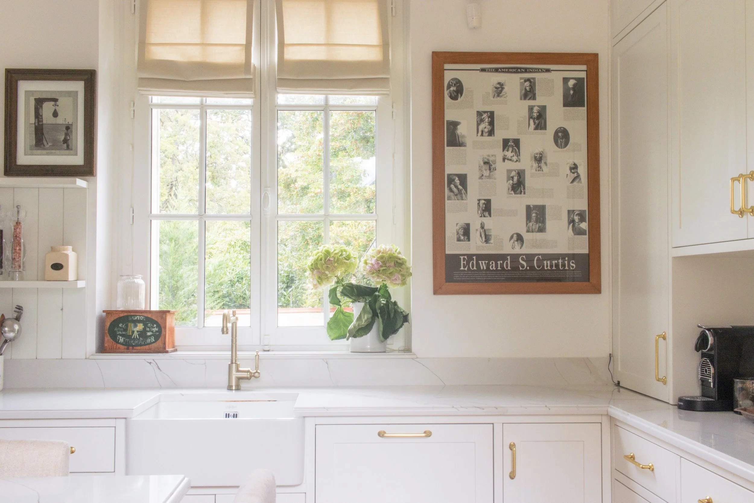 A bright kitchen with a large window, a white farmhouse sink, a plant with light green and pink flowers on the windowsill, and framed artwork on the wall, including a poster of Edward S. Curtis.