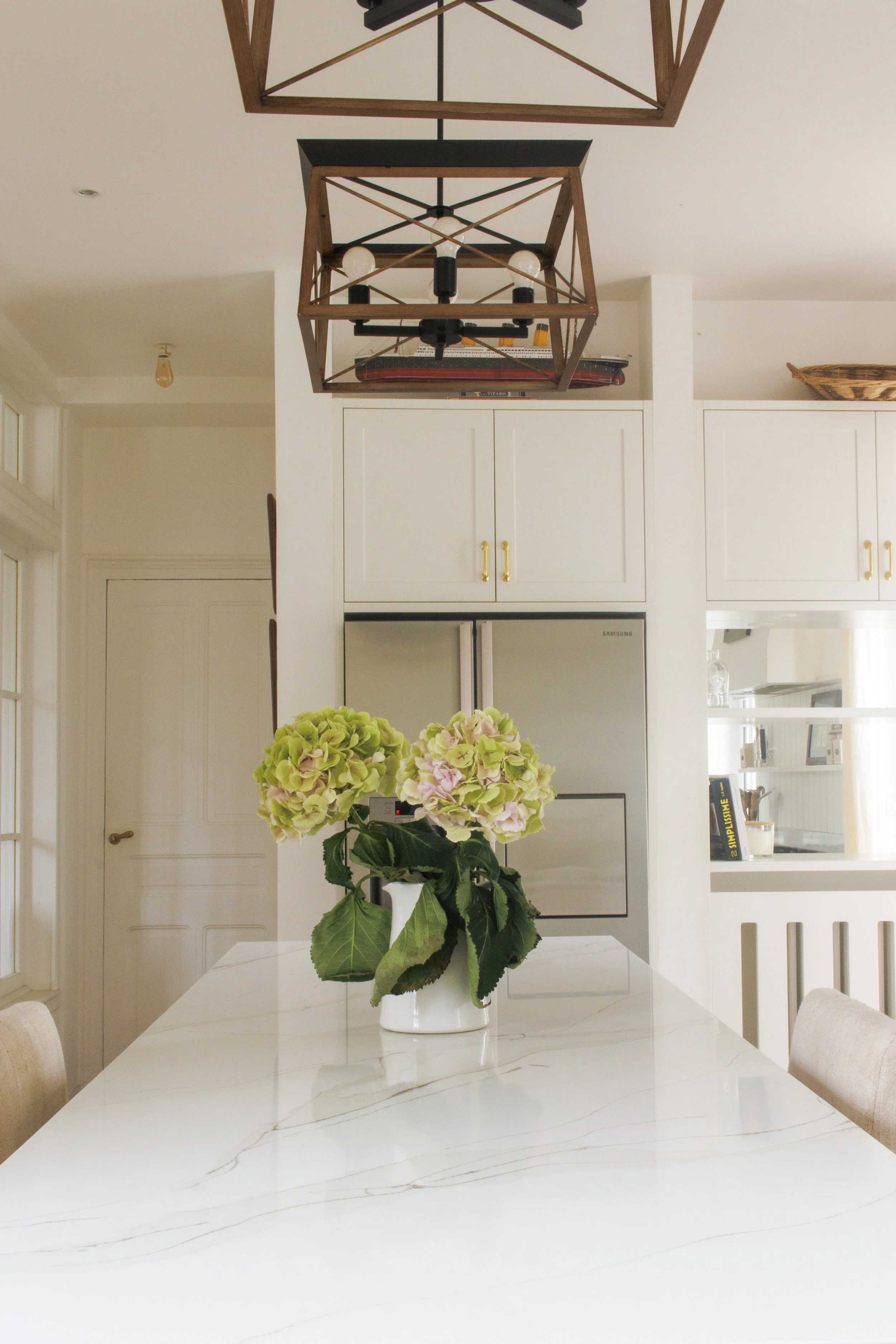 A dining room table with a white vase of pink and green hydrangeas in the center, a white table with a marble pattern, in a bright room with white walls, white cabinetry, and a refrigerator. A geometric wooden and black metal light fixture hangs from