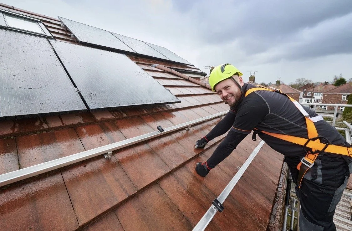 A man in safety gear, including a yellow helmet and harness, working on a sloped roof with solar panels and rain droplets, smiling at the camera.