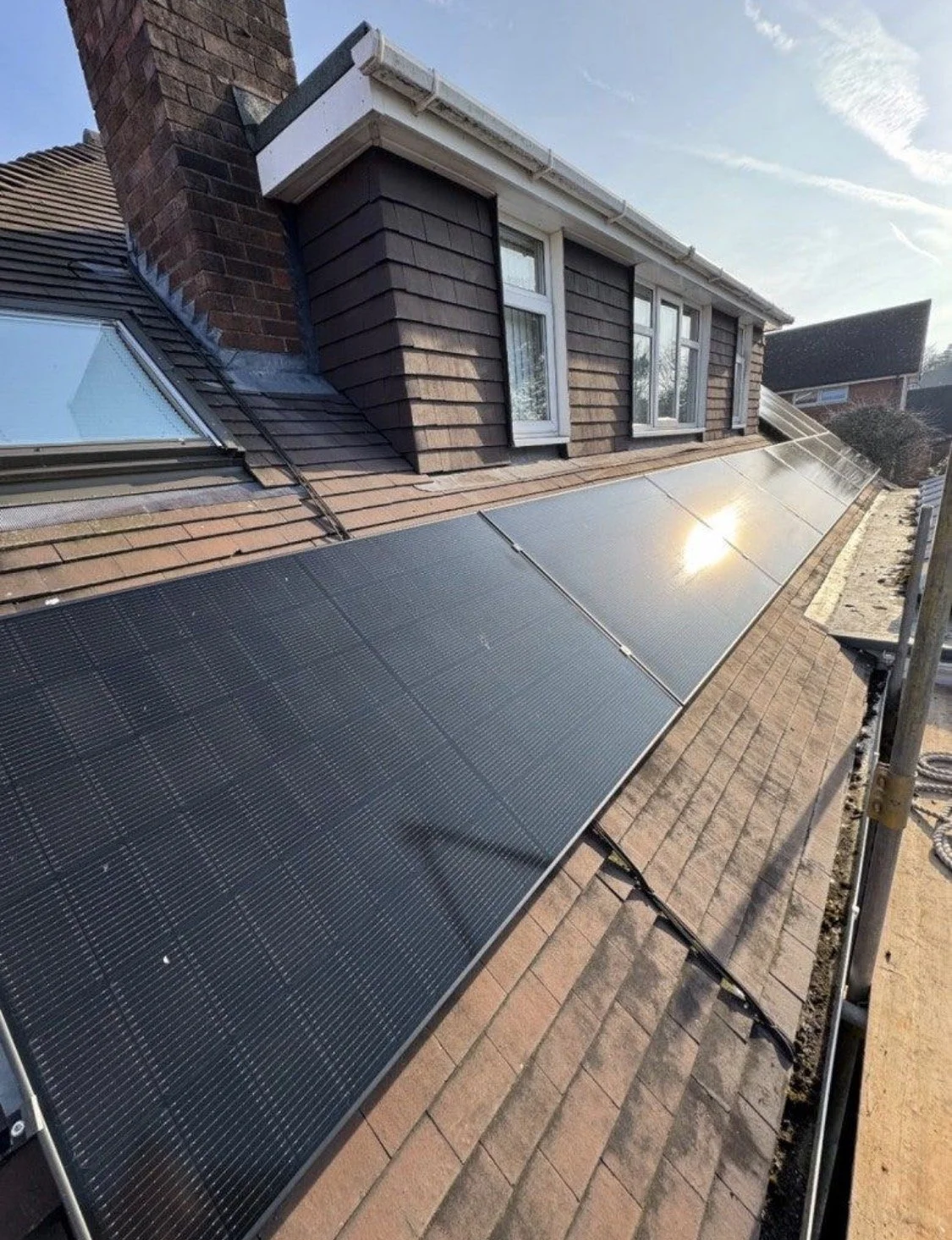 Solar panels installed on a sloped rooftop of a residential house, with a chimney and windows visible, under a clear blue sky.