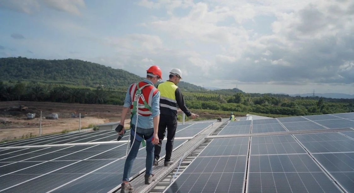 Three workers inspecting solar panels on a large solar farm under cloudy skies, with mountains and greenery in the background.