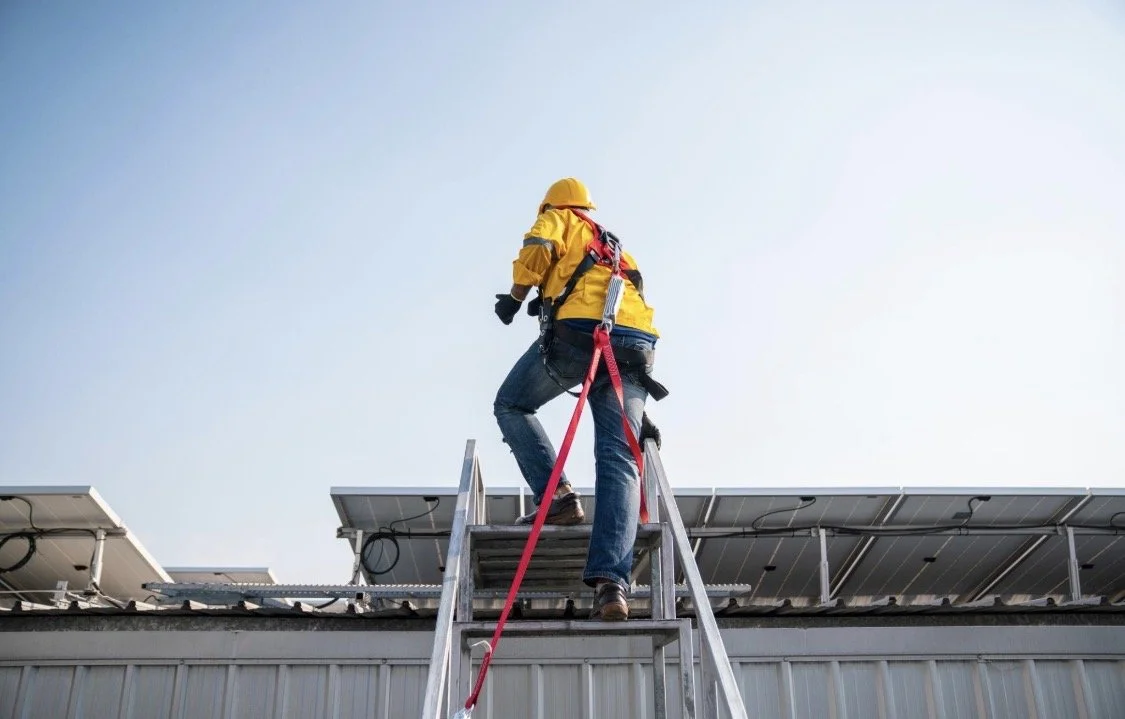 Worker in yellow safety gear and helmet climbing a ladder on a metal rooftop with solar panels.