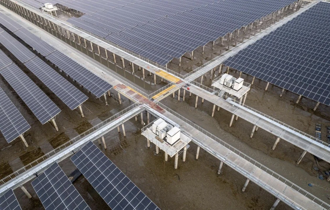 Aerial view of a solar farm with rows of solar panels mounted on elevated structures and connected walkways above barren land.