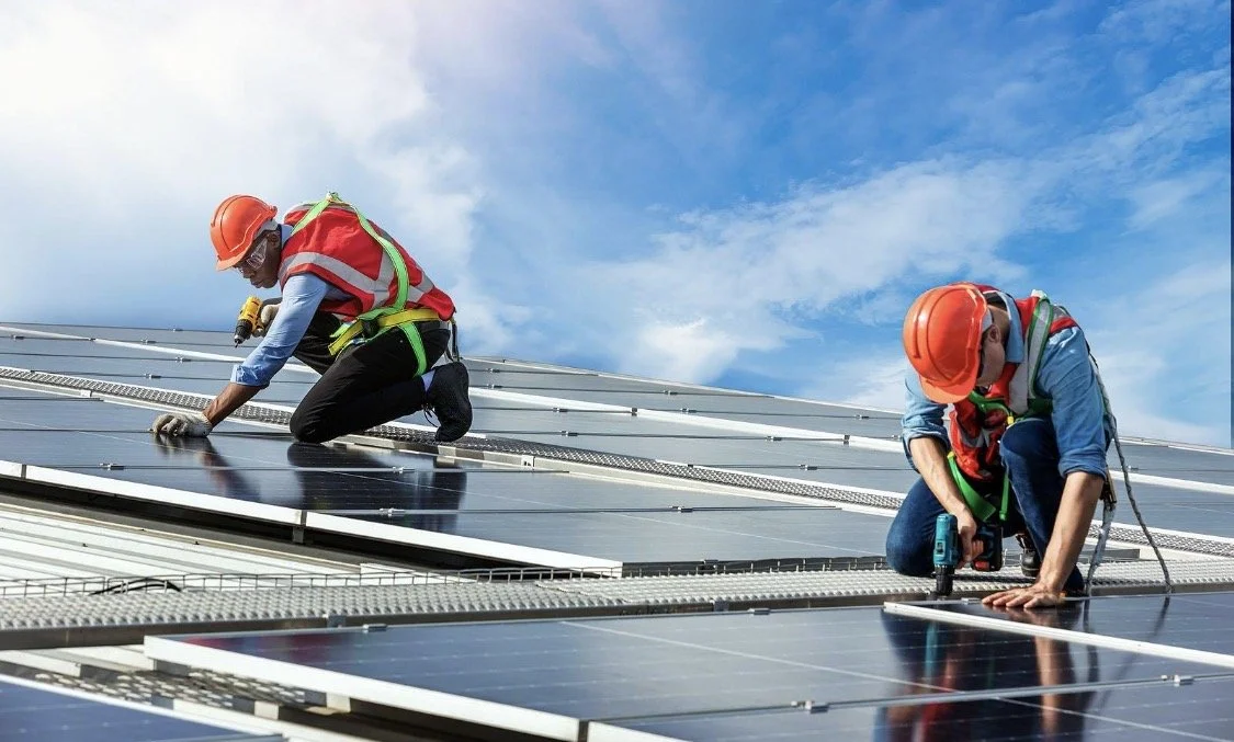 Two workers installing solar panels on a rooftop, both wearing orange safety helmets, vests, and kneeling while focusing on fixing the solar panels under a blue sky.