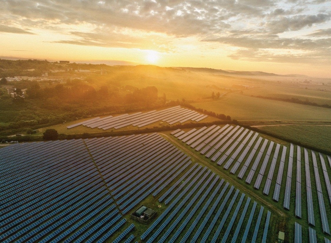 Aerial view of a large solar farm with rows of solar panels at sunset, surrounded by open fields and distant hills.
