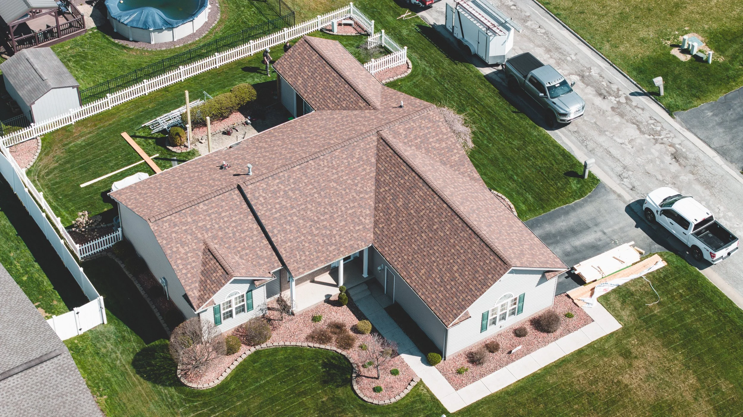 An aerial view of a suburban house with a brown tiled roof, a fenced backyard with a lawn, trees, and garden beds, and a driveway with parked trucks and cars.