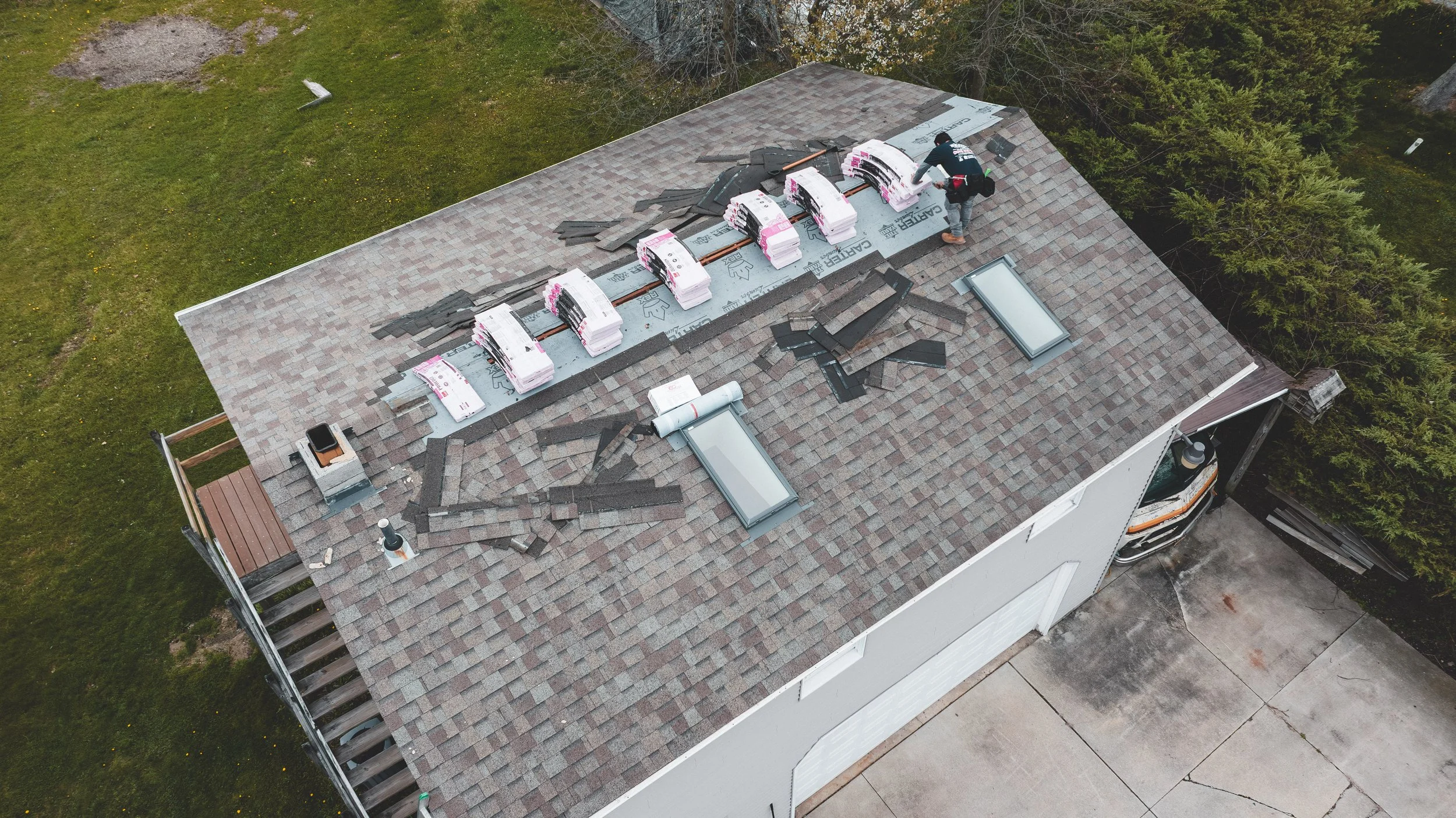 Aerial view of a house roof under construction with roofing materials and a worker installing new shingles, surrounded by green grass and trees.