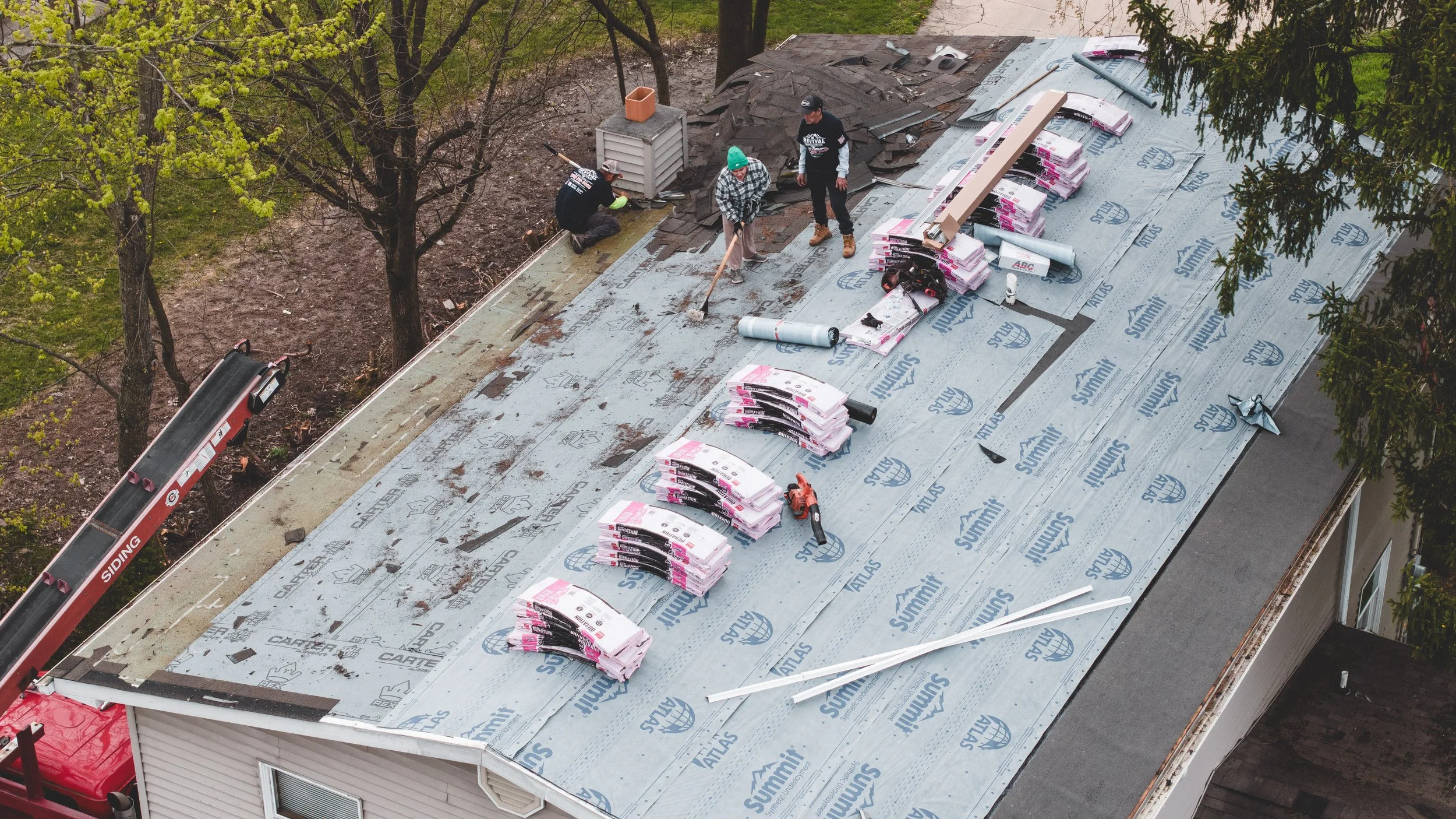 Workers installing roofing on a house using underlayment and shingles, with tools and materials laid out on the roof, and a ladder beside the building.