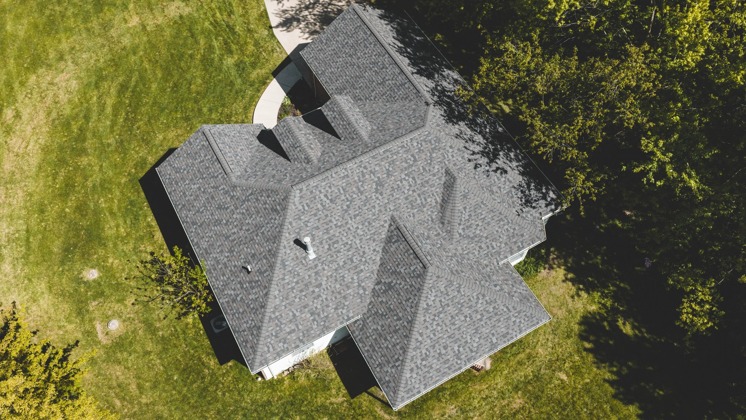 An aerial view of a house with a gray shingle roof surrounded by green grass and trees.