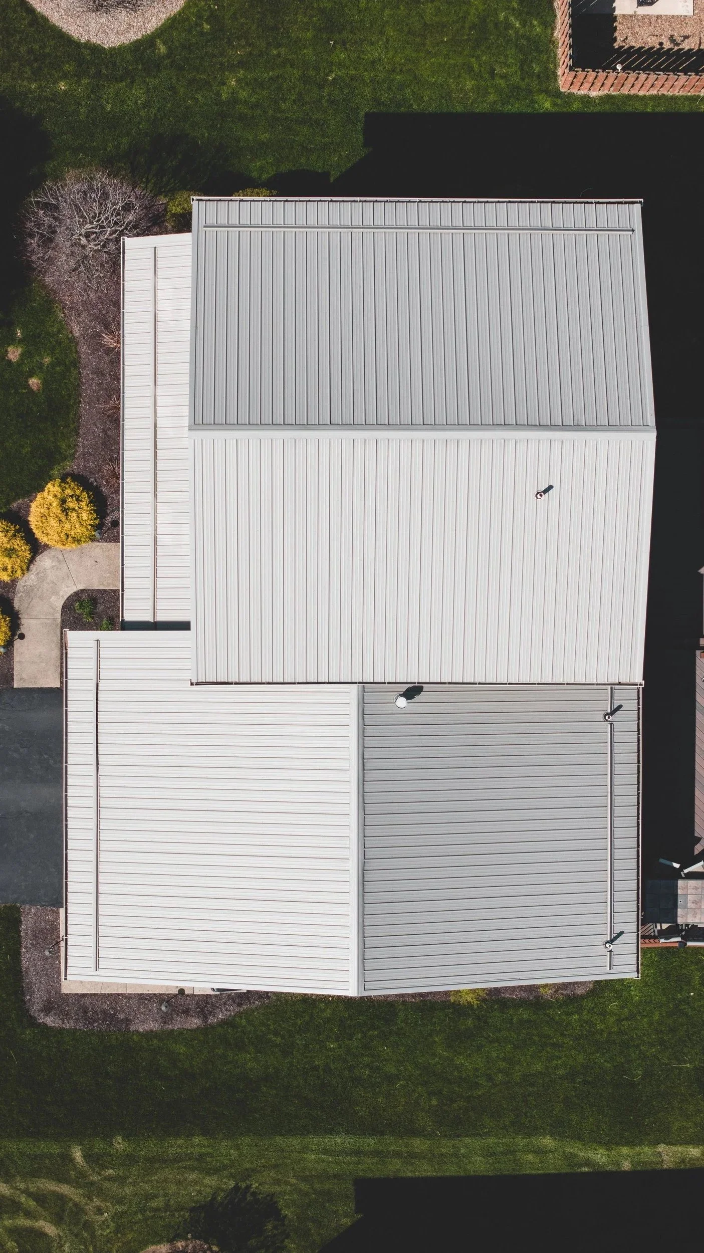 Aerial view of three metal-roofed buildings with varying shades of gray, surrounded by green lawns, trees, and pathways.