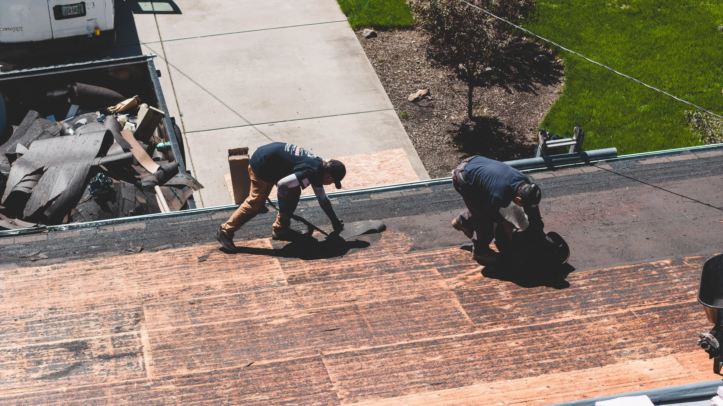 Two workers are remodeling a roof, one is removing old shingles while the other is installing new shingles, with a backyard lawn and sidewalk visible in the background.