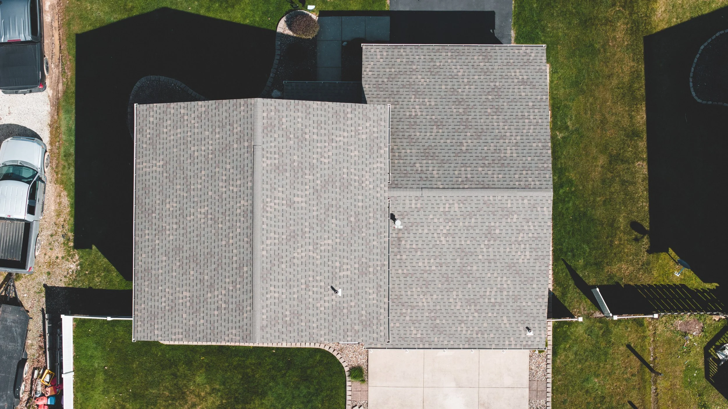 An aerial view of a house with a gray shingled roof, a driveway, parked cars, green lawn, and surrounding fences.